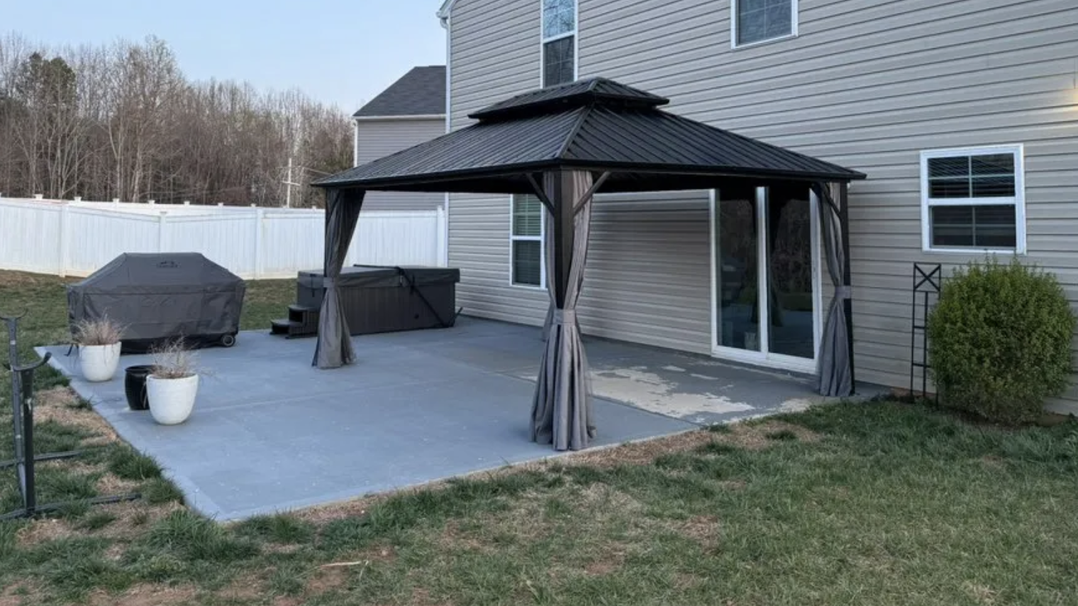 Backyard patio with a gazebo, hot tub, grill covered with a tarp, and potted plants, enclosed by a white fence with a house in the background.