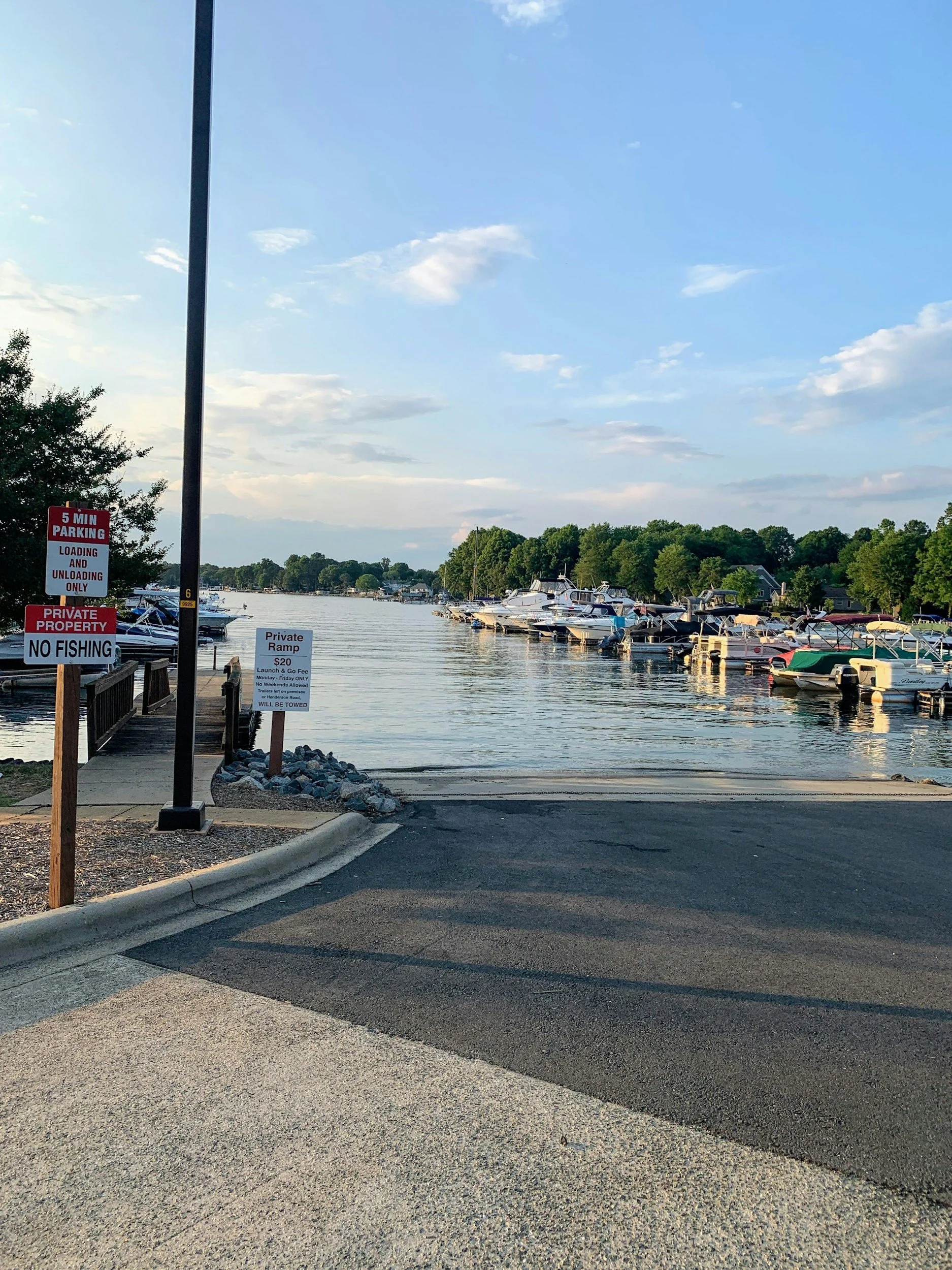 A marina with boats docked along the water, with signs indicating parking restrictions and a private ramp, under a partly cloudy sky.