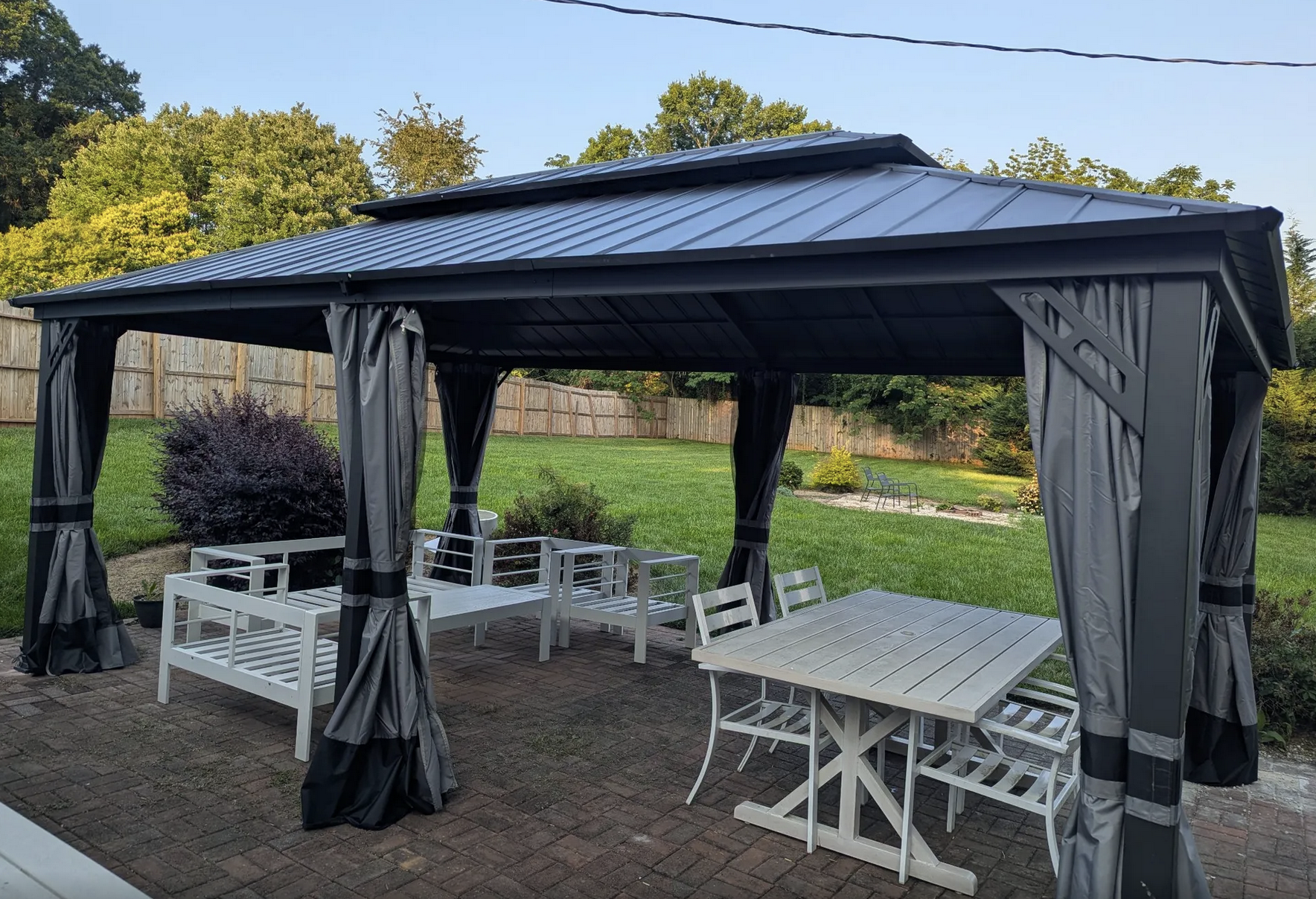 A backyard patio with a black metal gazebo, white outdoor furniture including a table and chairs, brick flooring, and greenery with trees and bushes in the background.