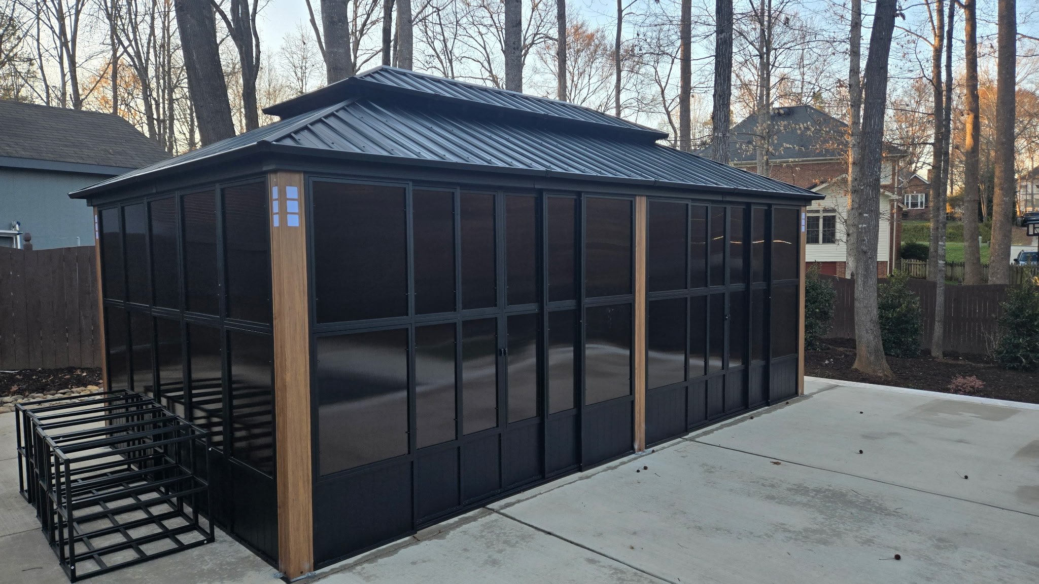 Black framed screened-in outdoor structure with wooden accents and metal roof, situated on a concrete patio, surrounded by a backyard with trees and neighboring houses.