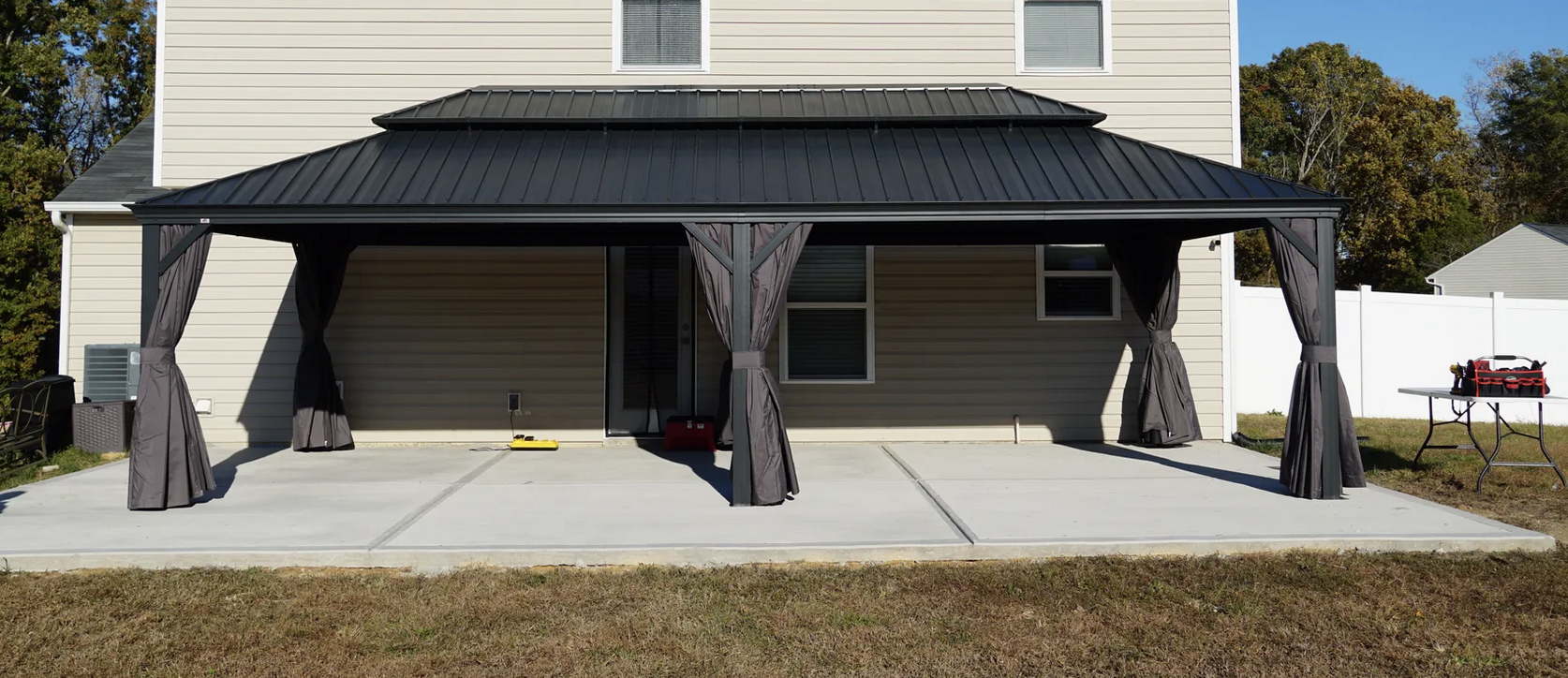 Backyard patio with a black metal roof porch, supported by four posts with curtains. The concrete patio has two spaces, one with a red toolbox, and there is a table with a black and red toolbox on the right side. The house has beige siding and two windows.