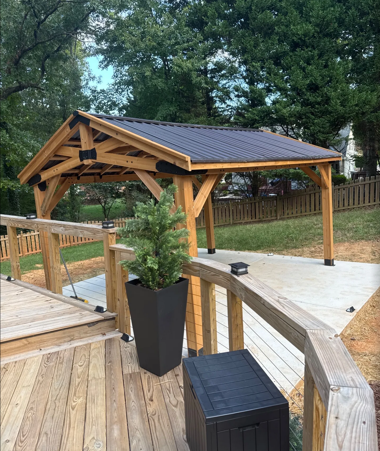 Wooden deck with a railing and a potted evergreen plant, leading to a concrete area with a wooden shelter supported by wooden beams, and a black storage box on the deck. Trees and a fence are in the background.
