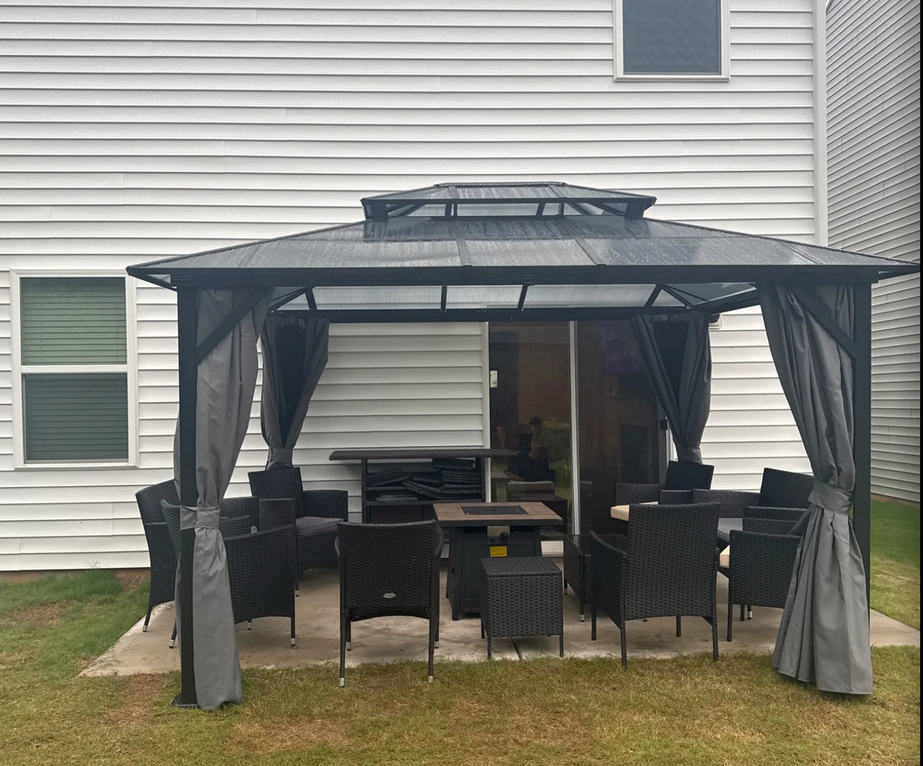 Outdoor patio furniture set under a black metal gazebo with curtains, on a concrete patio next to a white house with beige siding.