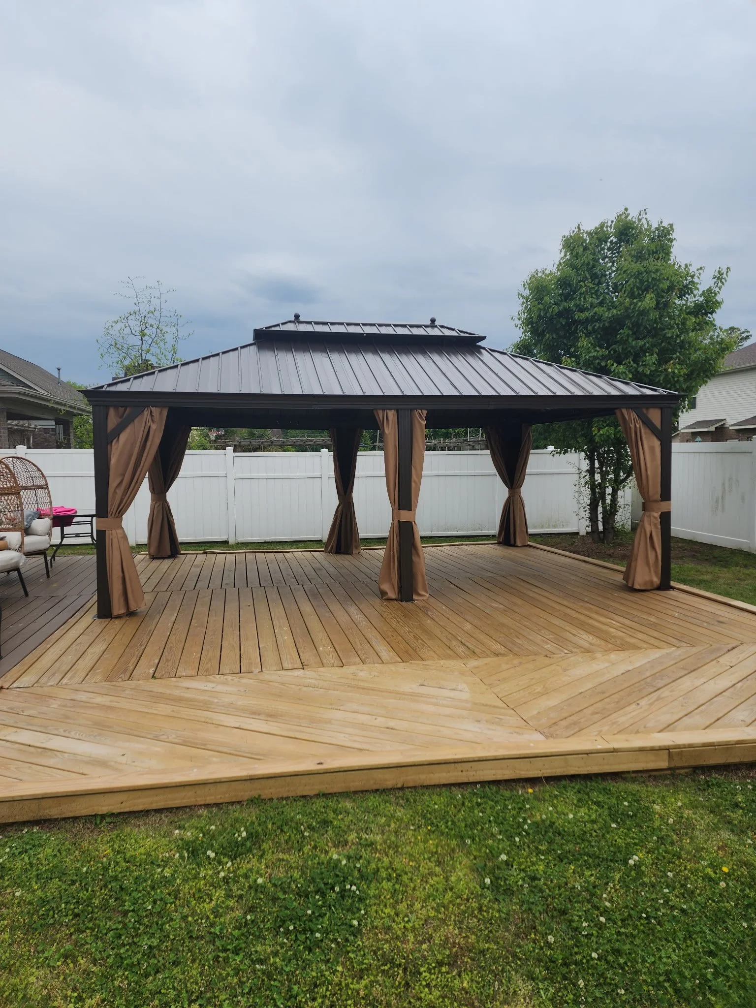 Wooden deck with a black gazebo with curtains in a backyard, white fences, trees, and cloudy sky.