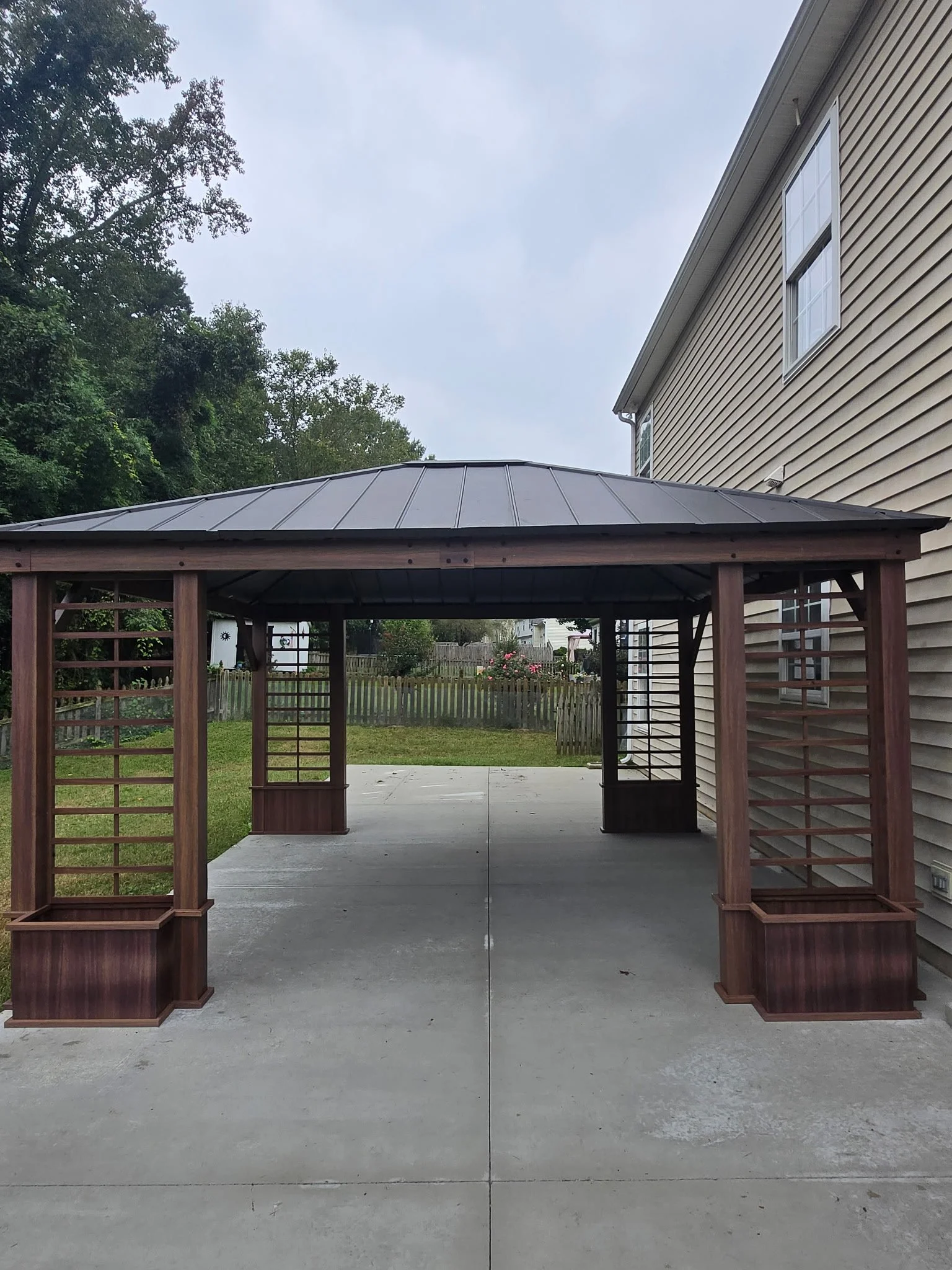 A wooden gazebo with a metal roof over a concrete driveway, adjacent to a beige house with siding, in a backyard with lawn, trees, and a wooden picket fence.