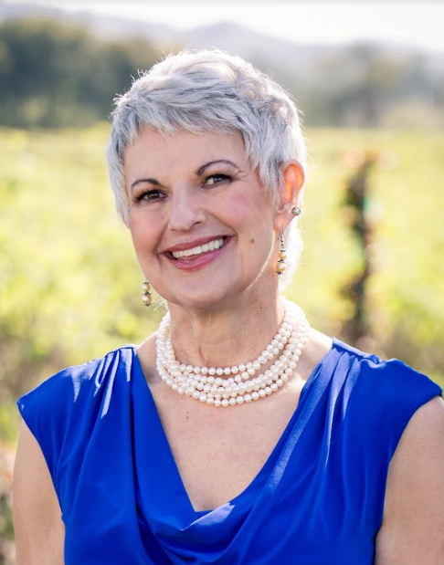 Portrait of Doreen, with silver hair in a beautiful blue dress and a multi-string of white pearls, smiling widely and looking towards the camera.
