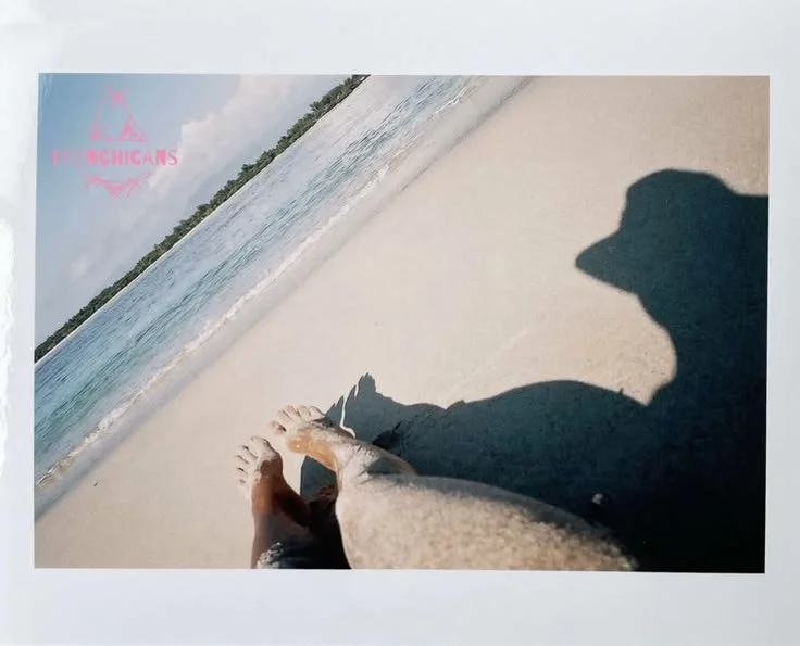 A person sitting on a sandy beach, near the ocean, with a distant shoreline and cloudy sky in the background.