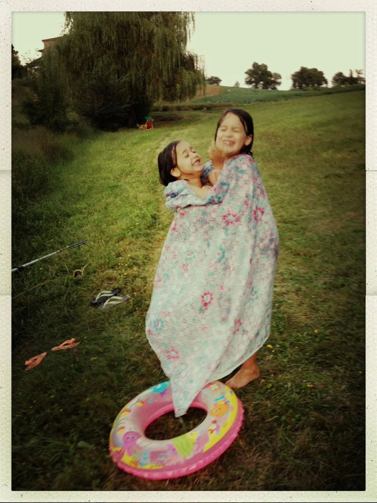 Two young girls smiling and hugging, wrapped in a colorful blanket, standing outdoors on grass near a small pool, with trees and a hill in the background.