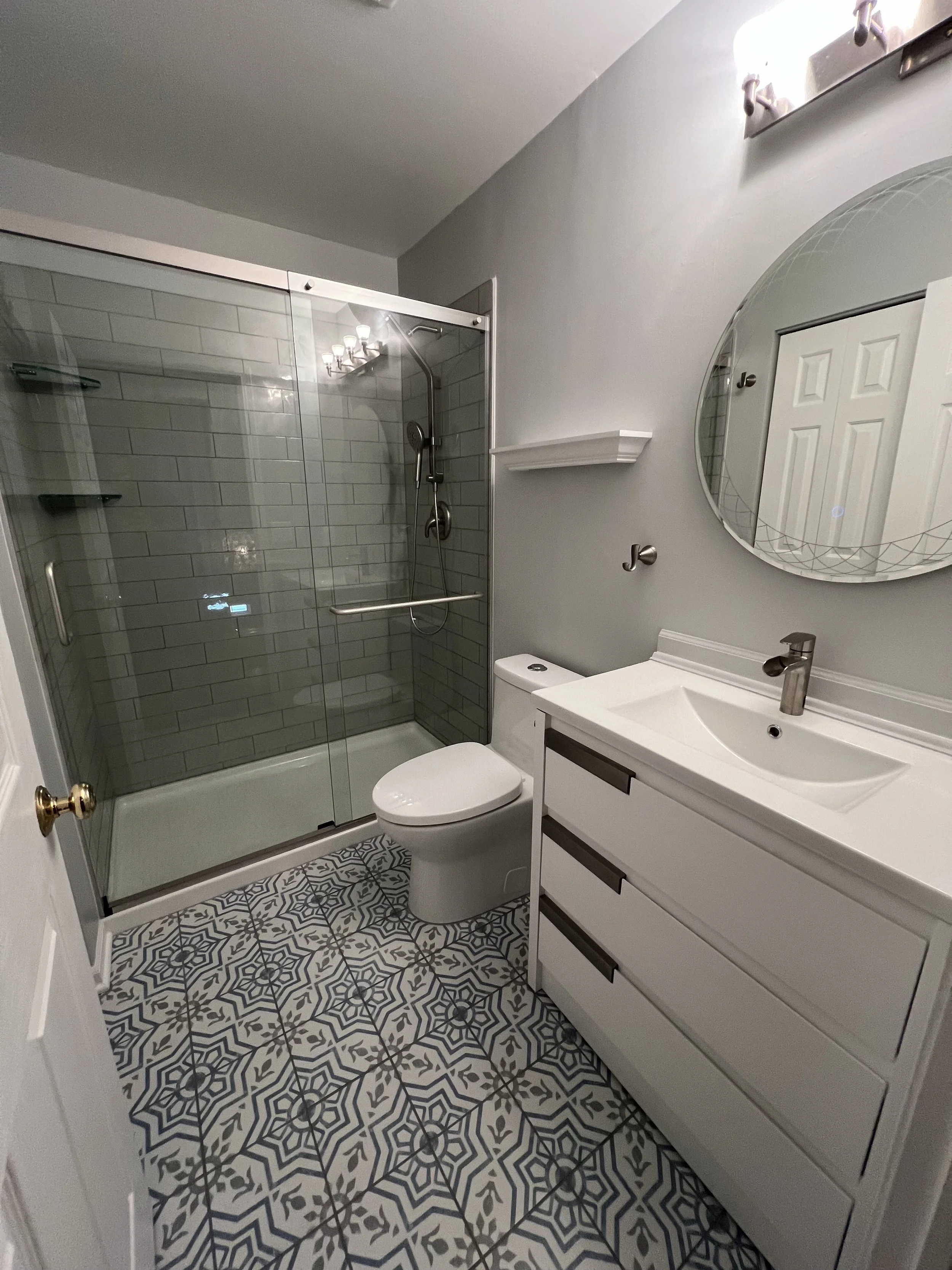 Bathroom with a shower enclosure, a white toilet, a vanity with a sink, a round mirror, and patterned tile flooring.