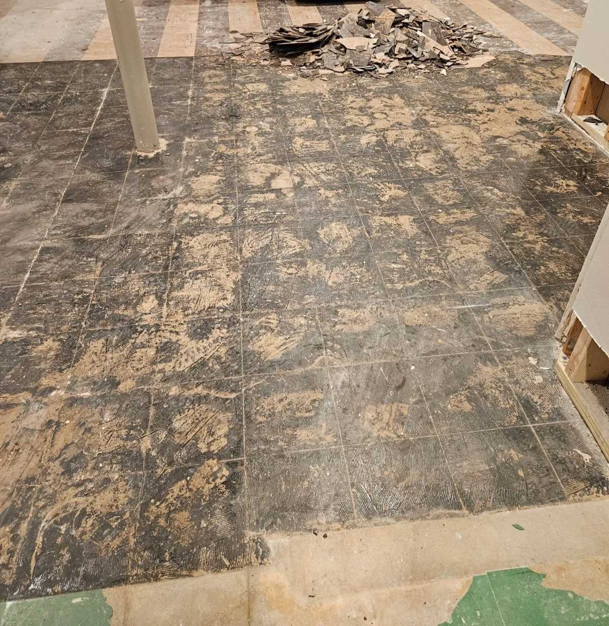 Floor with black tiles covered in construction dust and debris, with torn up green and beige flooring at the edges and some drywall in the background.