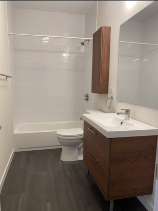 Bathroom with white bathtub, toilet, wooden vanity with sink, mirror, and a wooden cabinet above the sink.