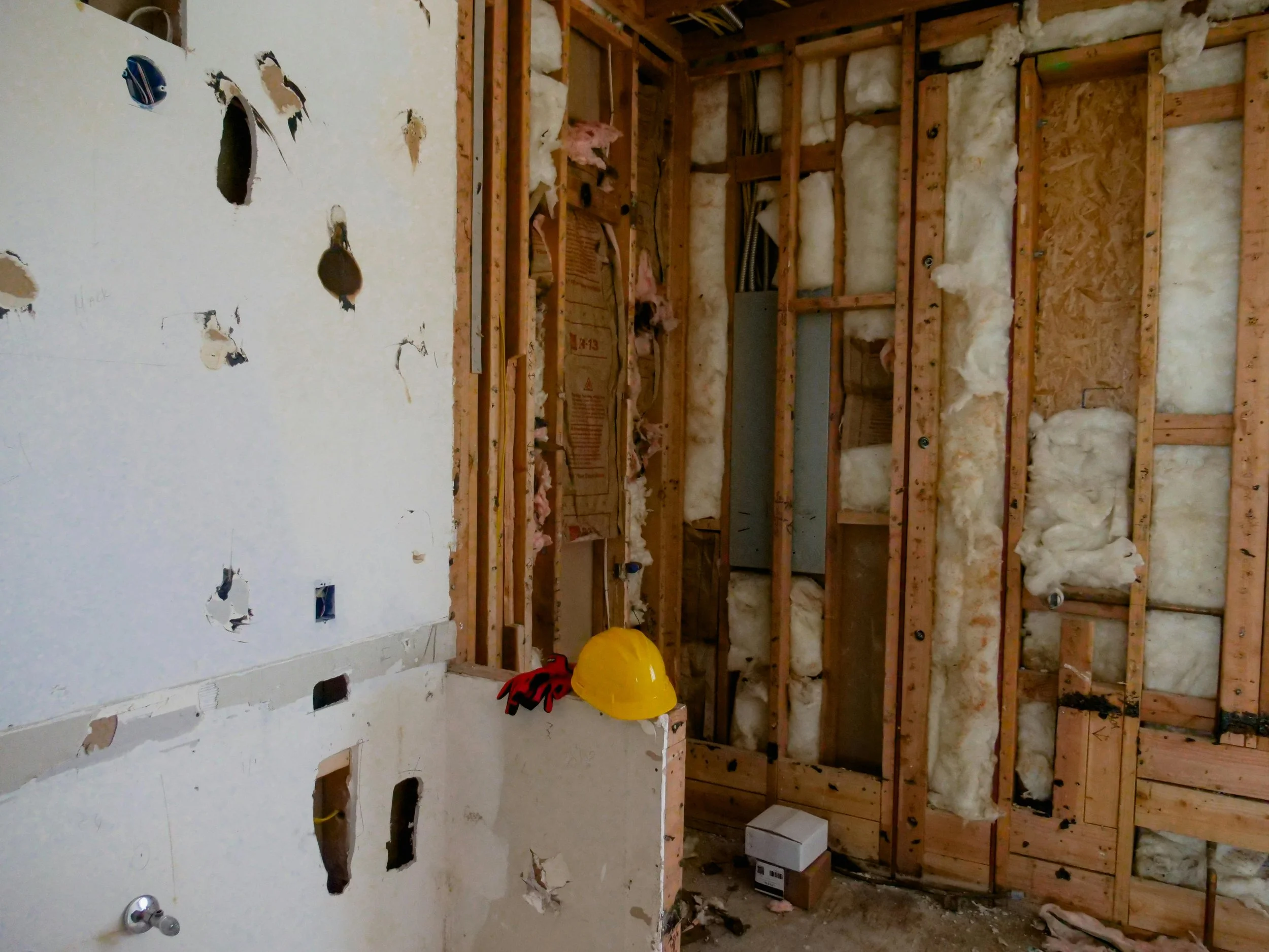 Interior view of a room under construction with exposed wooden studs, insulation, and partially demolished drywall. Safety helmet and gloves on a ledge in the foreground.