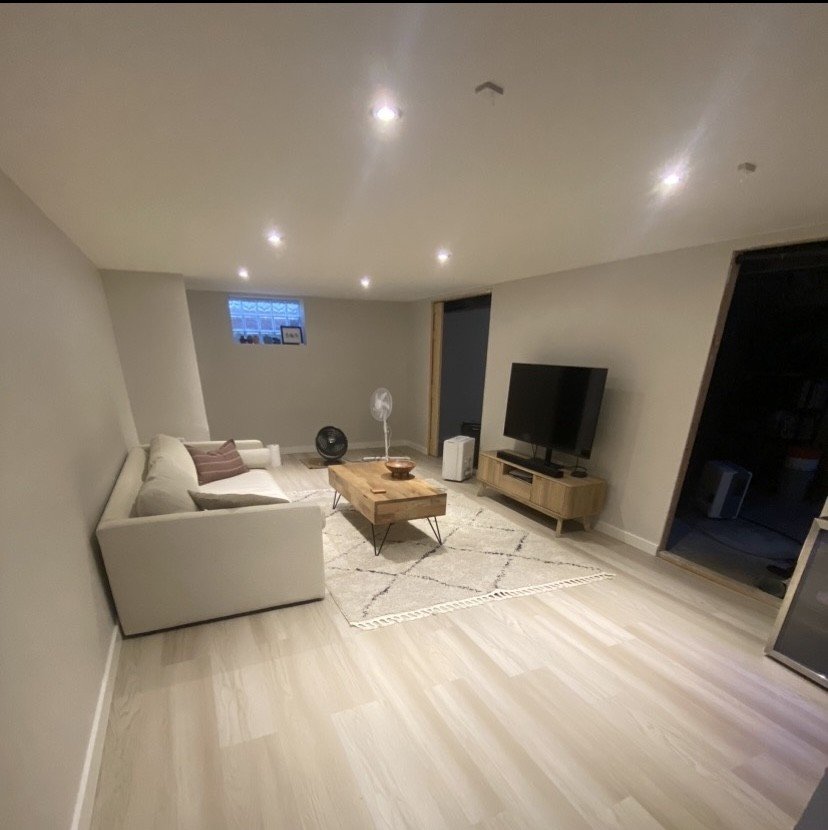A living room with a beige sofa, wooden coffee table, flat-screen TV on a wooden stand, and a small window. The room has recessed ceiling lights and a light-colored wood floor.