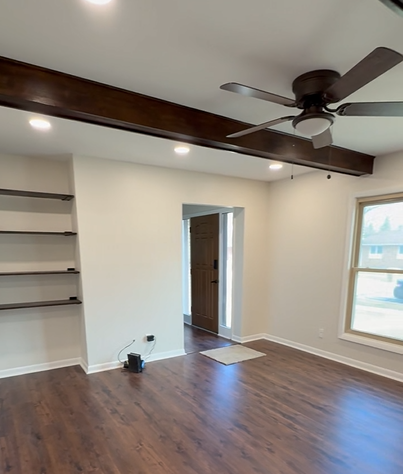 Empty living room with white walls, hardwood floors, a ceiling fan, with a doorway leading to another room and a large window letting in natural light.