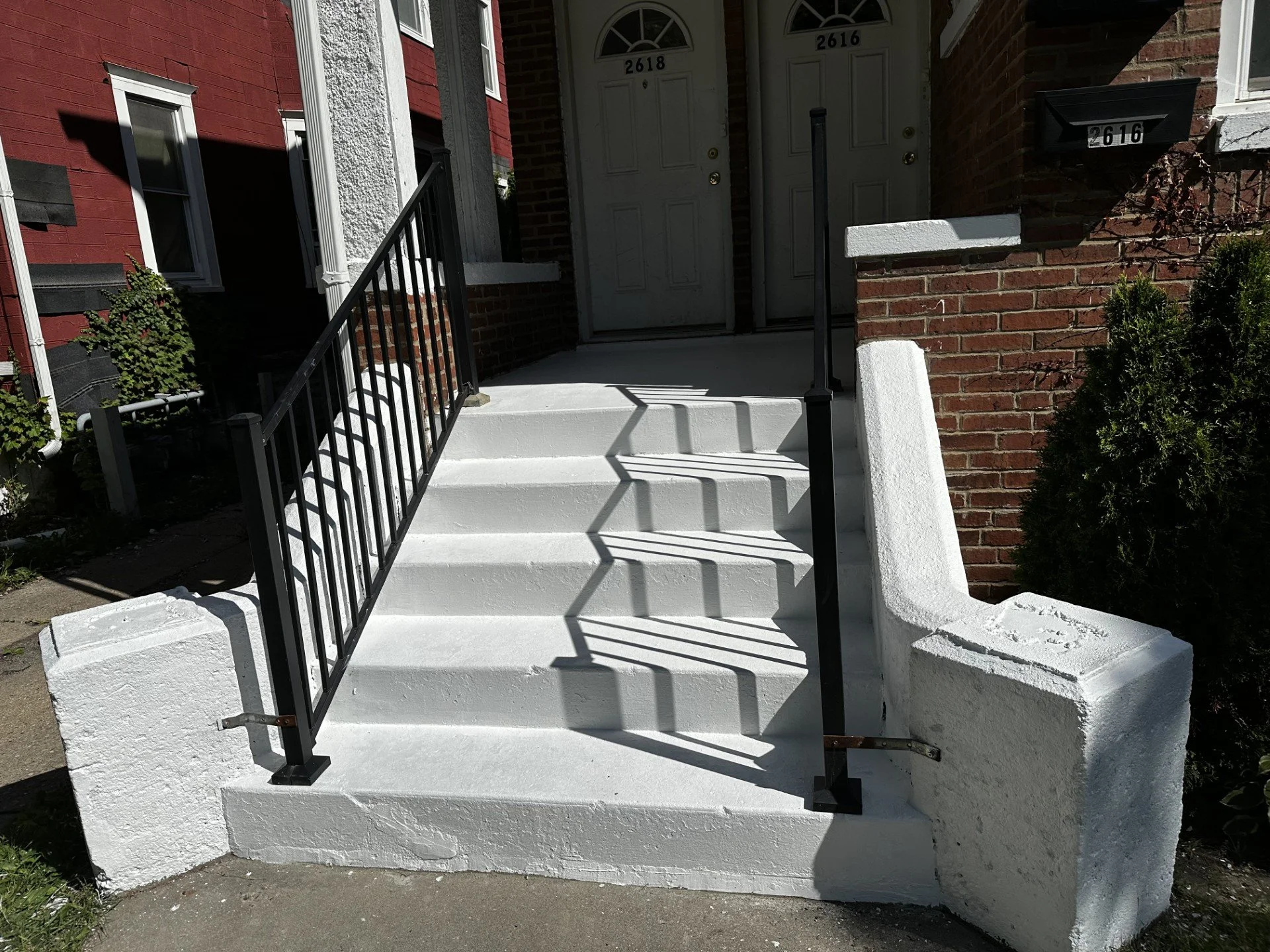 Concrete stairs leading to a front door with black metal railing on the left side, double white doors at the top, brick exterior walls, and a small shrub on the right side.