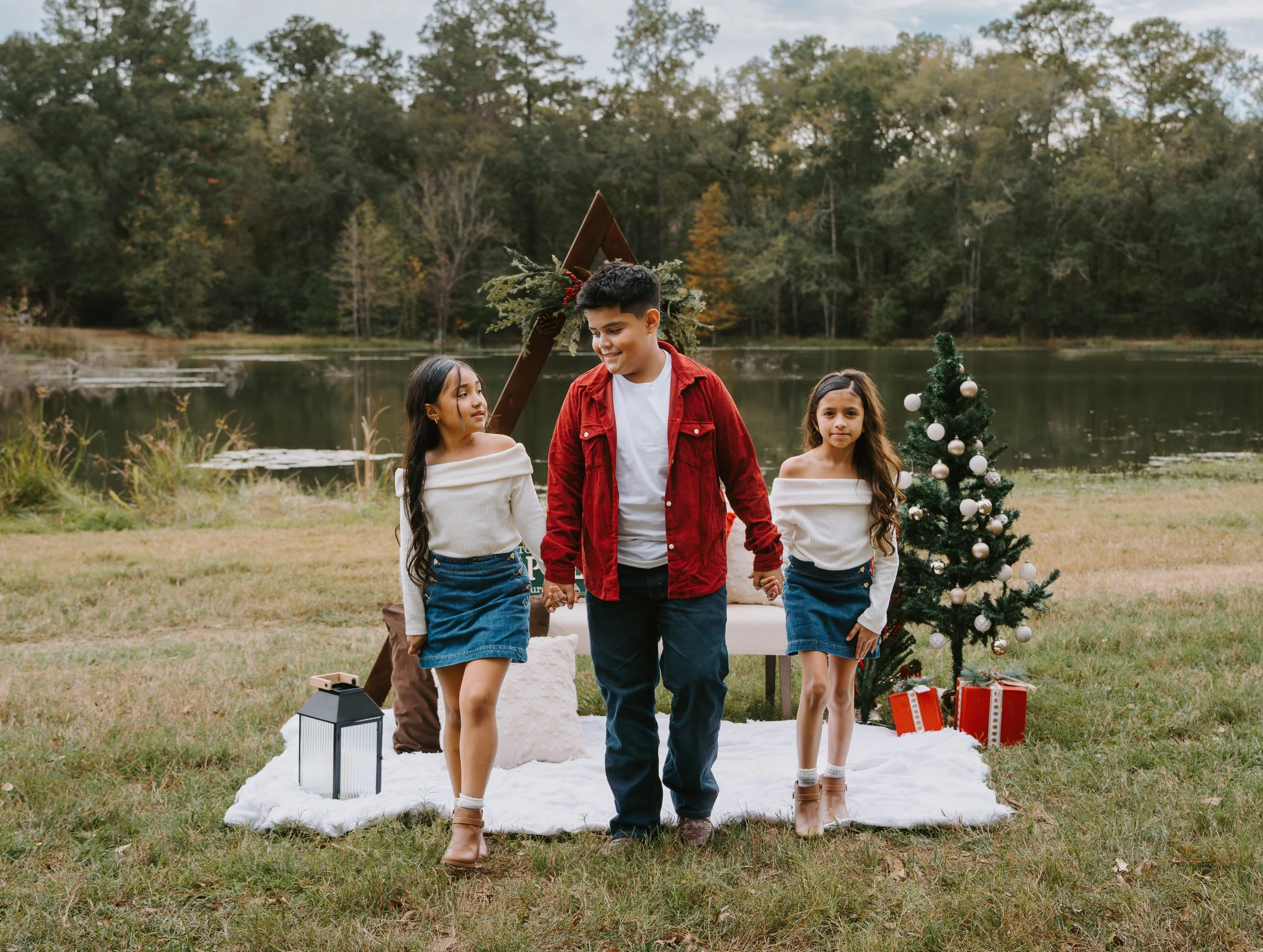 A family with two young girls and a teenage boy outdoors by a lake, holding hands and walking on grass during daytime. The background features trees, a small decorated Christmas tree with ornaments, a white blanket, and some wrapped gifts.
