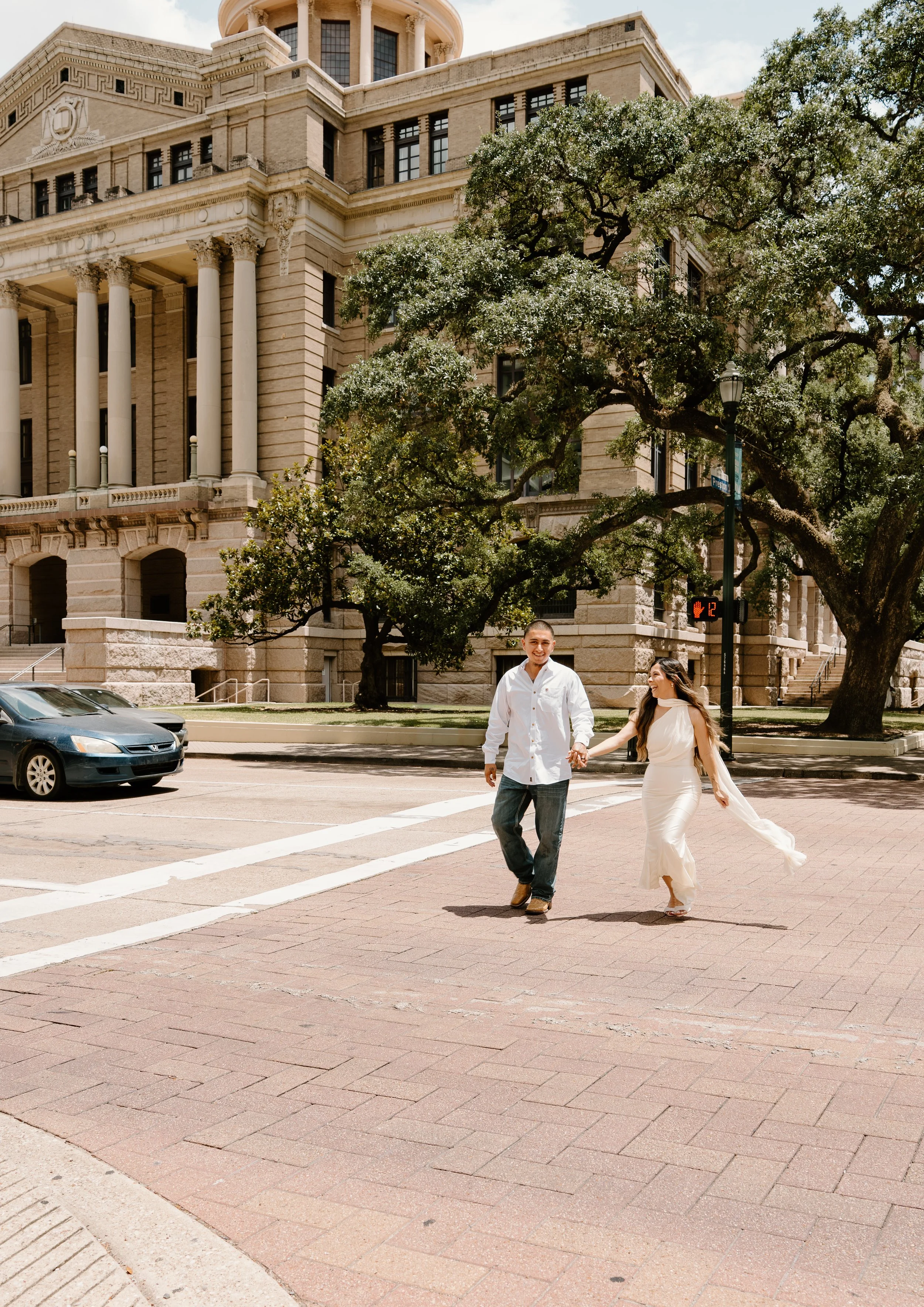 A smiling man and woman holding hands and crossing a city street in front of a large historic building with columns and trees.