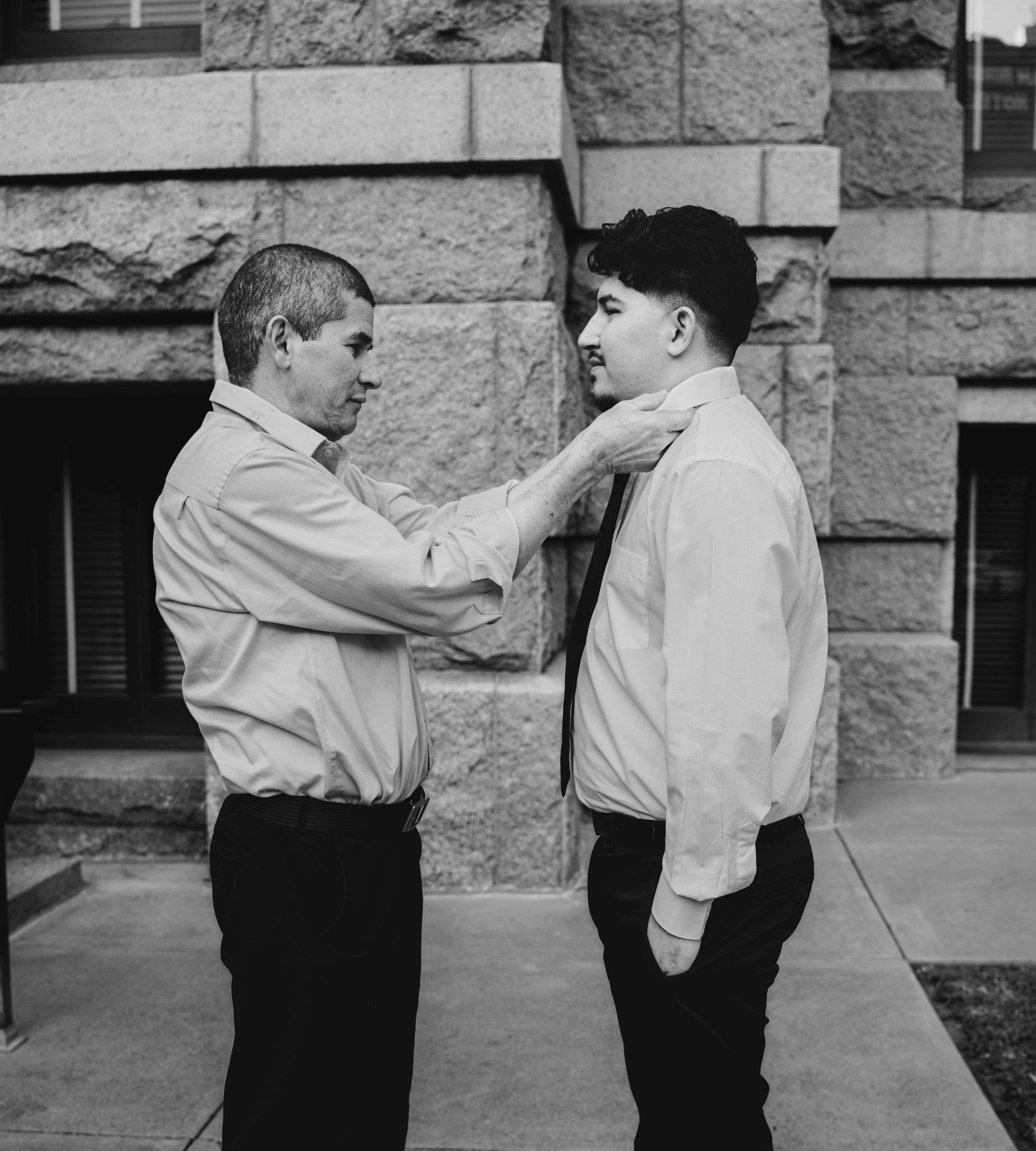 A Father helps his son adjust his tie outside of a building with stone walls before his wedding.