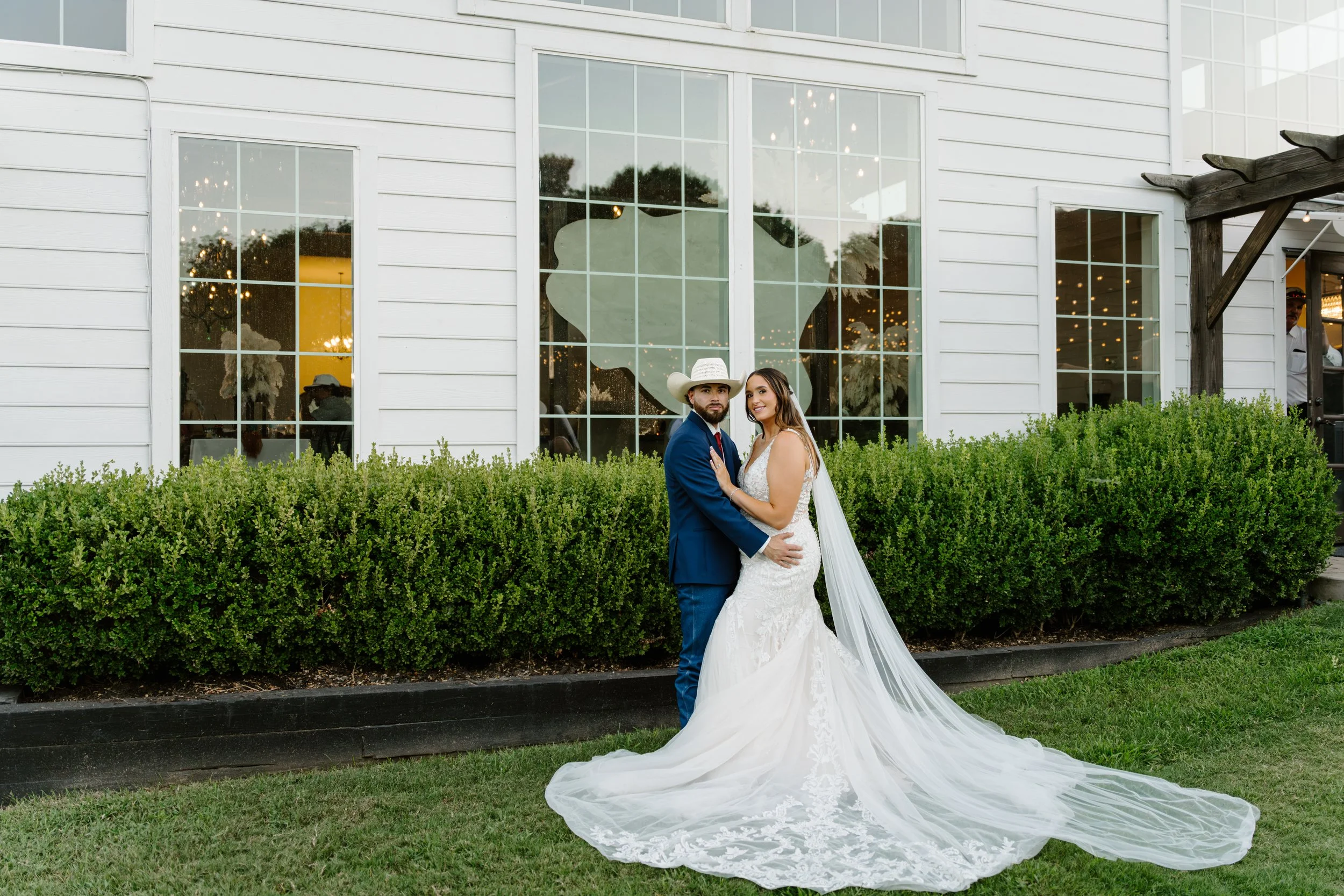 A newlywed couple stands outside a white building with large windows, surrounded by greenery. The groom wears a navy suit and a cowboy hat, while the bride wears a white lace wedding dress with a long train and a veil. They pose closely together, hol