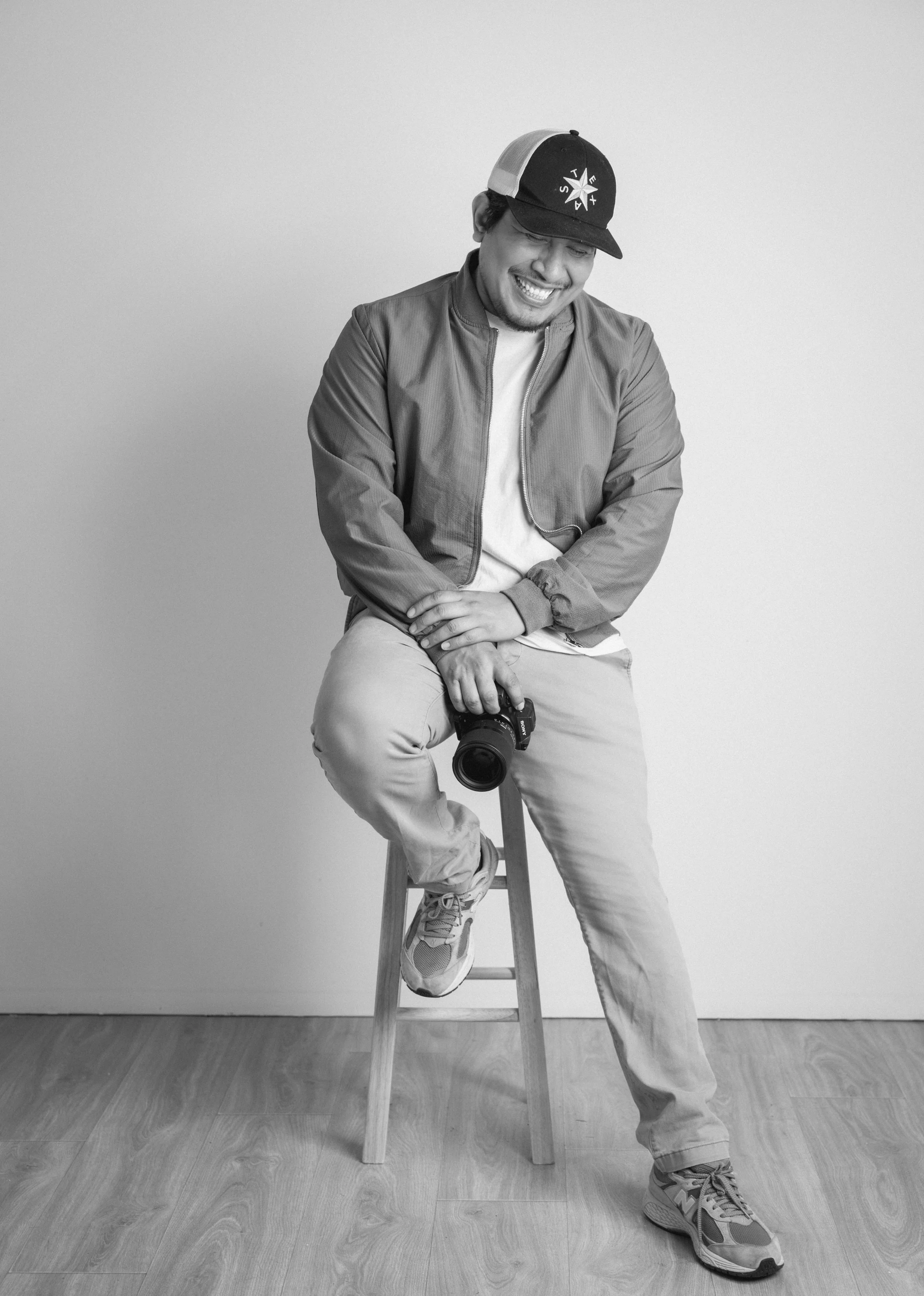 Black and white photo of a man sitting on a wooden stool, smiling with eyes closed, holding a camera in his right hand, wearing a cap, a jacket, and sneakers, against a plain wall.
