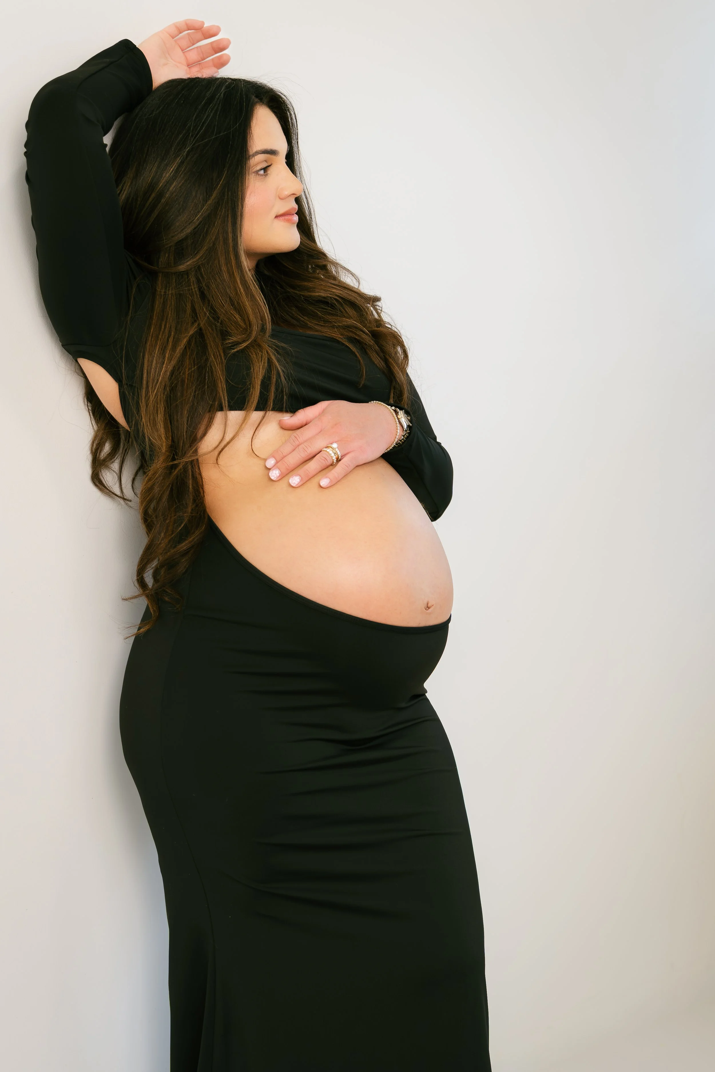 A pregnant woman with long brown hair leaning against a white wall, wearing a black dress and jewelry, with her eyes closed and arm raised above her head.