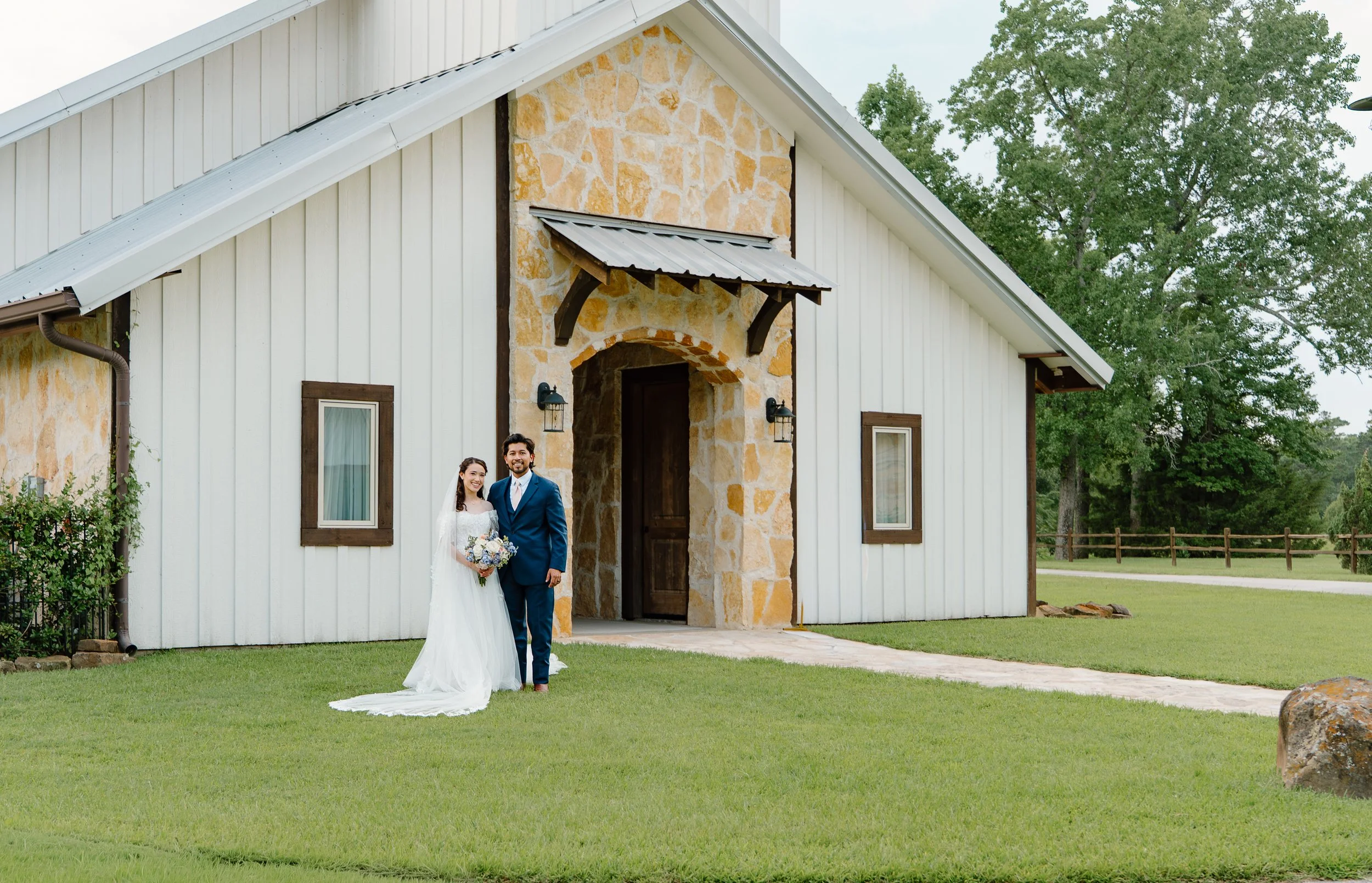 A bride and groom standing outside a house with a stone and white wood exterior, green lawn, and trees in the background.