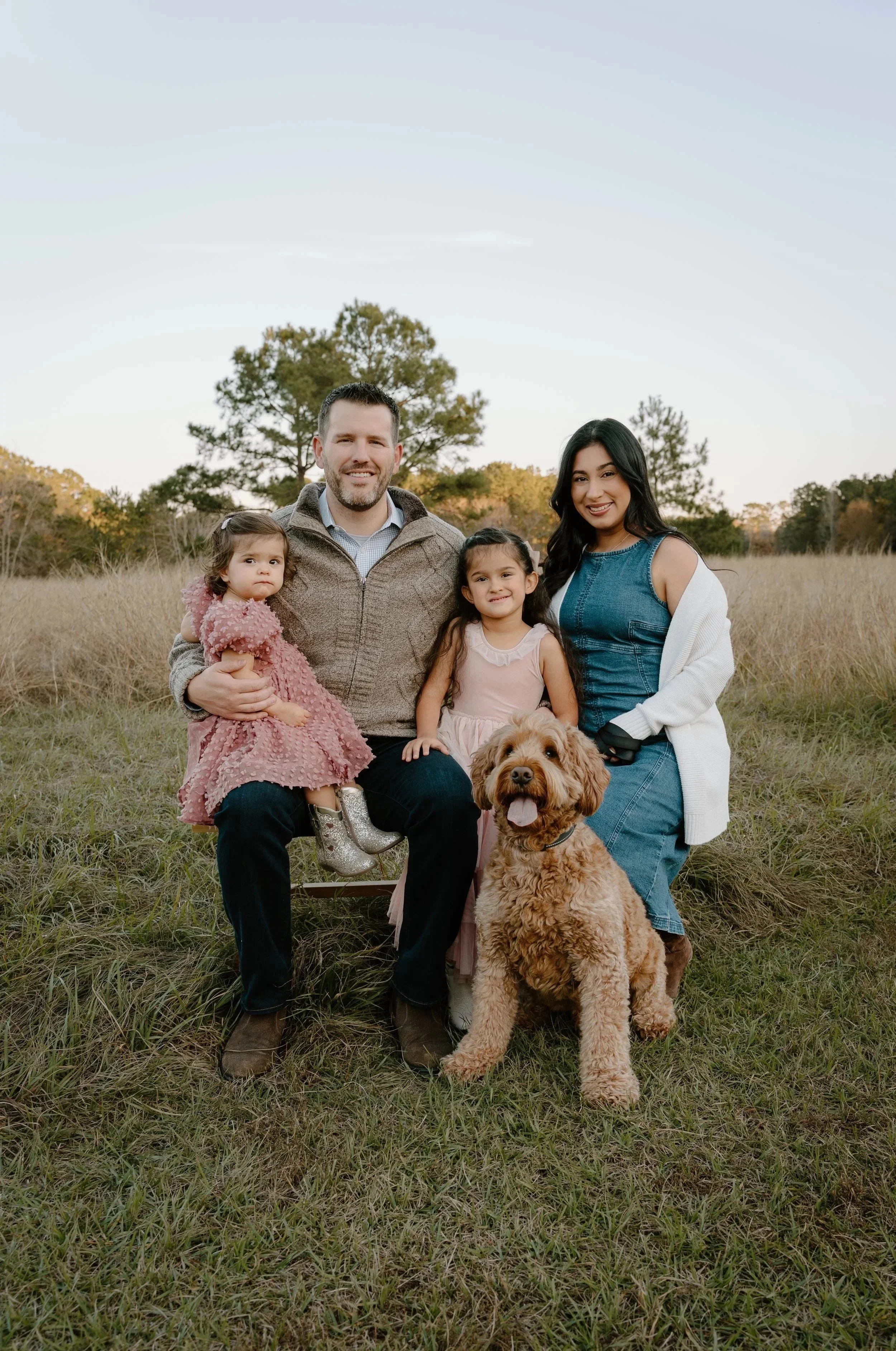 A family of four with two young girls and a woman with an adorable brown curly-haired dog, sitting on a grassy field during sunset.
