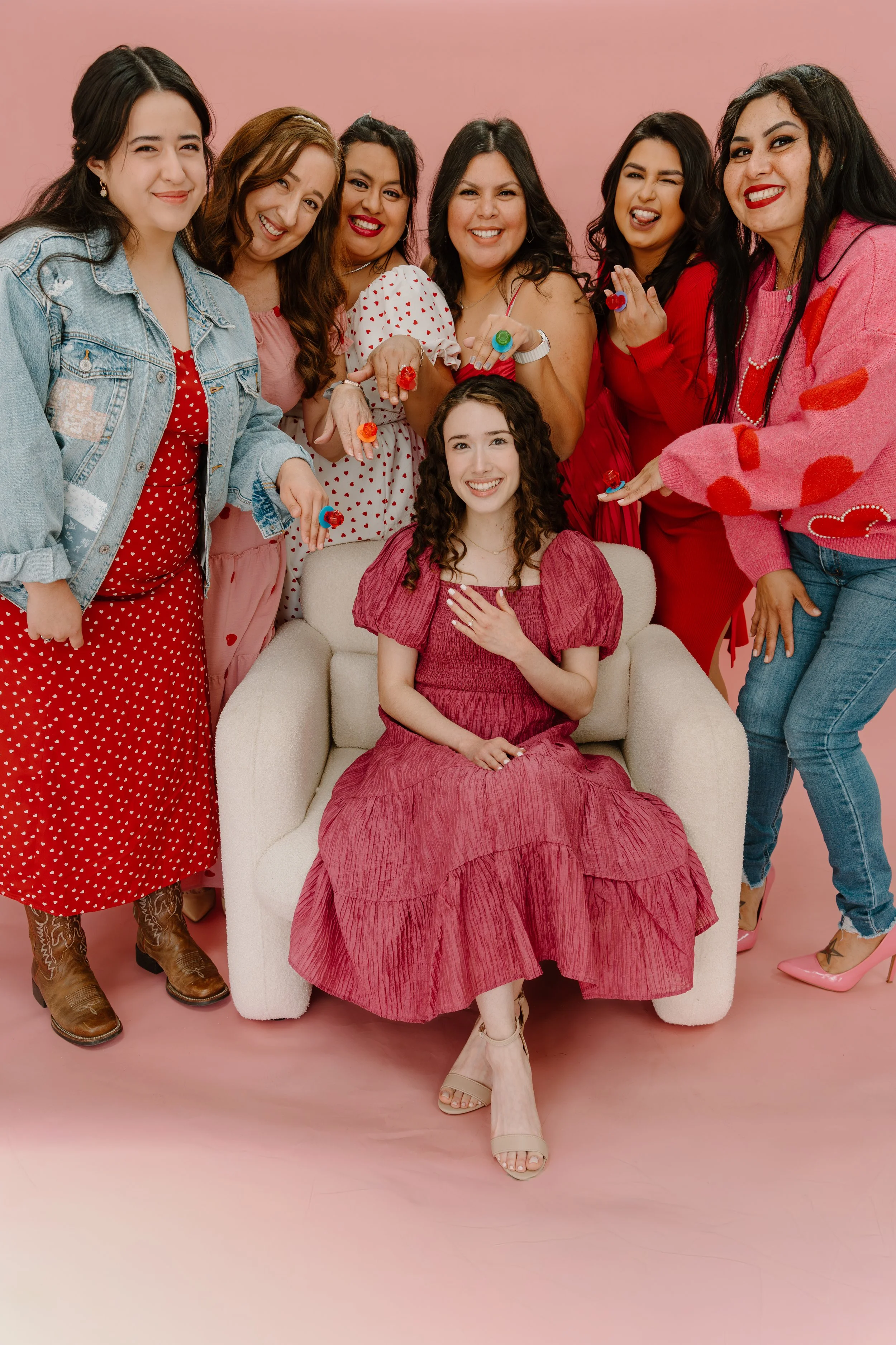 A young woman in a red dress sitting on a white armchair surrounded by seven women pointing at her with smiles, against a pink background.