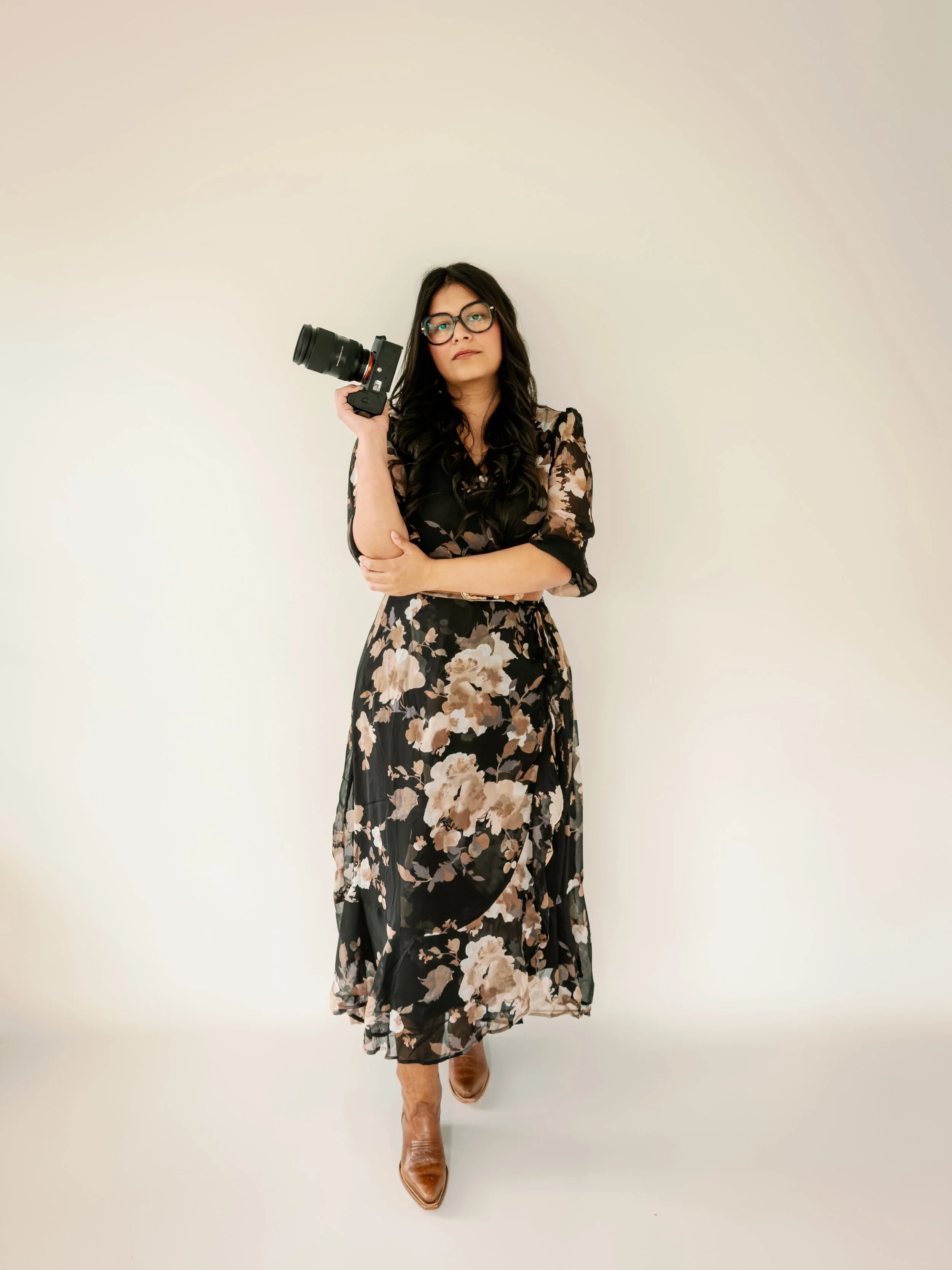 Woman in glasses holding a camera, wearing a floral dress and cowboy boots, standing against a plain beige wall.