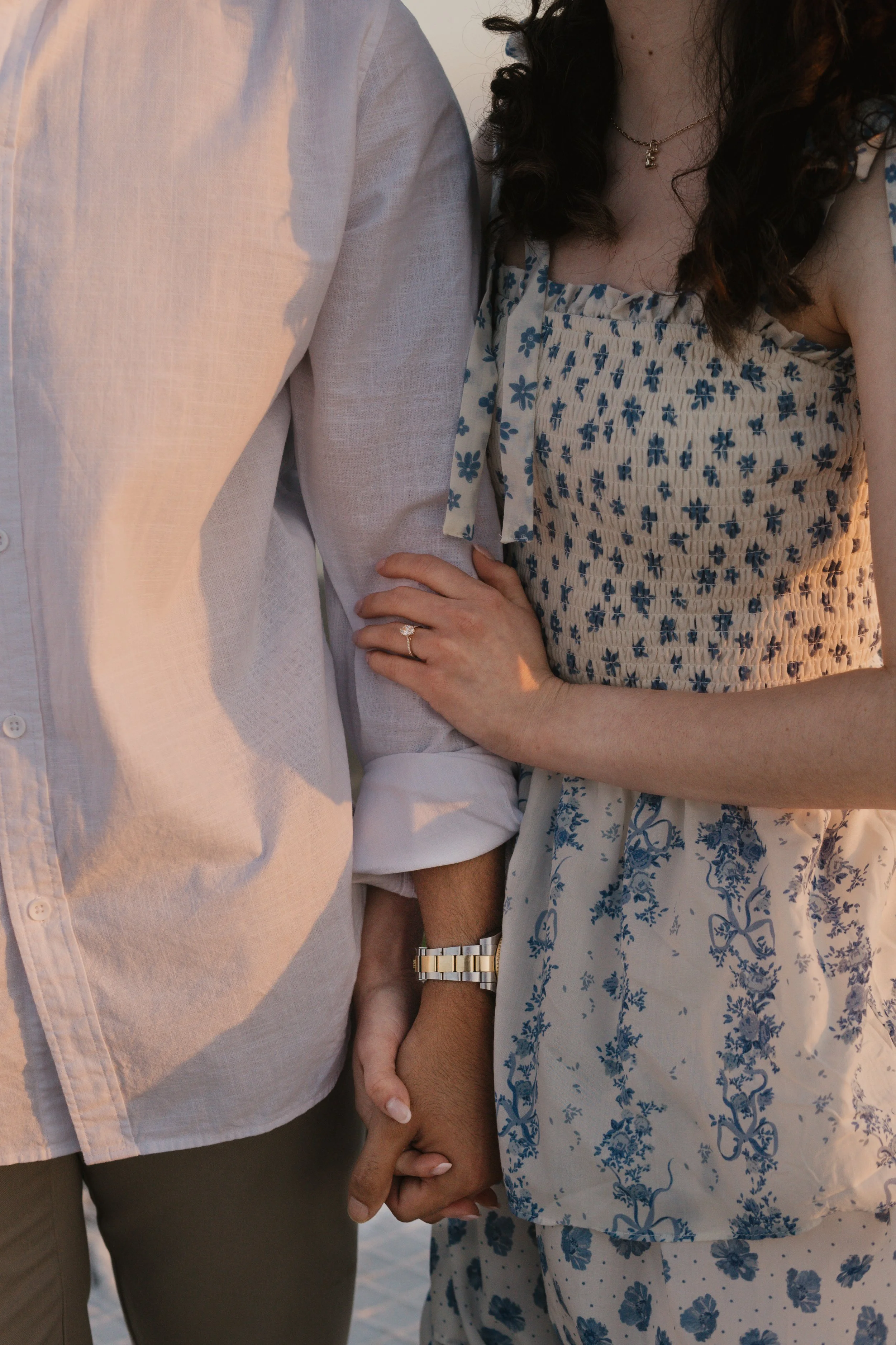 A close-up of a couple holding hands, with the woman wearing a ring and a blue and white floral dress, and the man wearing a white shirt and a watch. The woman rests her hand on the man's arm.