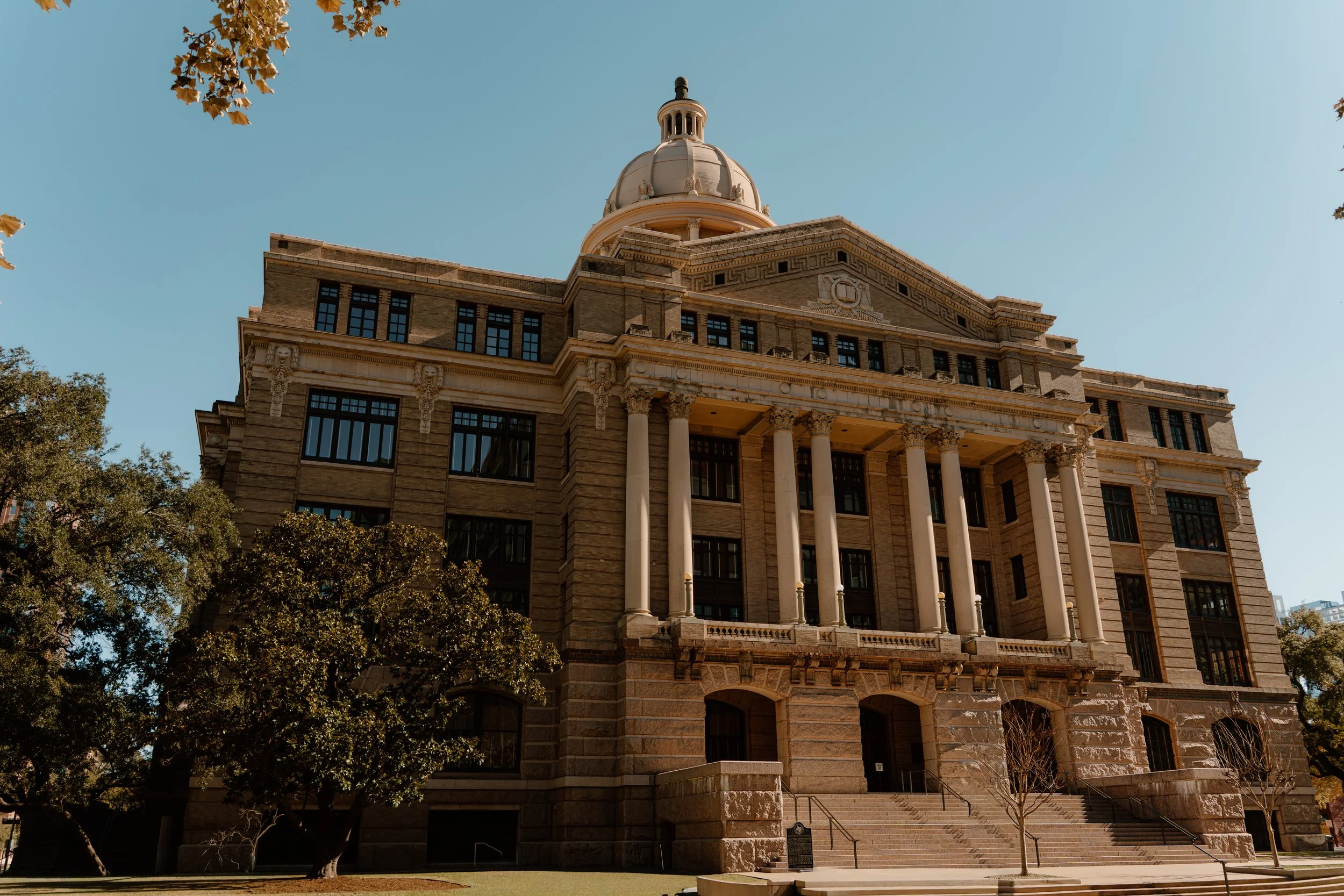 A historic government building with classical architecture, large columns, steps leading to the entrance, and a dome on top, set against a clear blue sky with trees around.