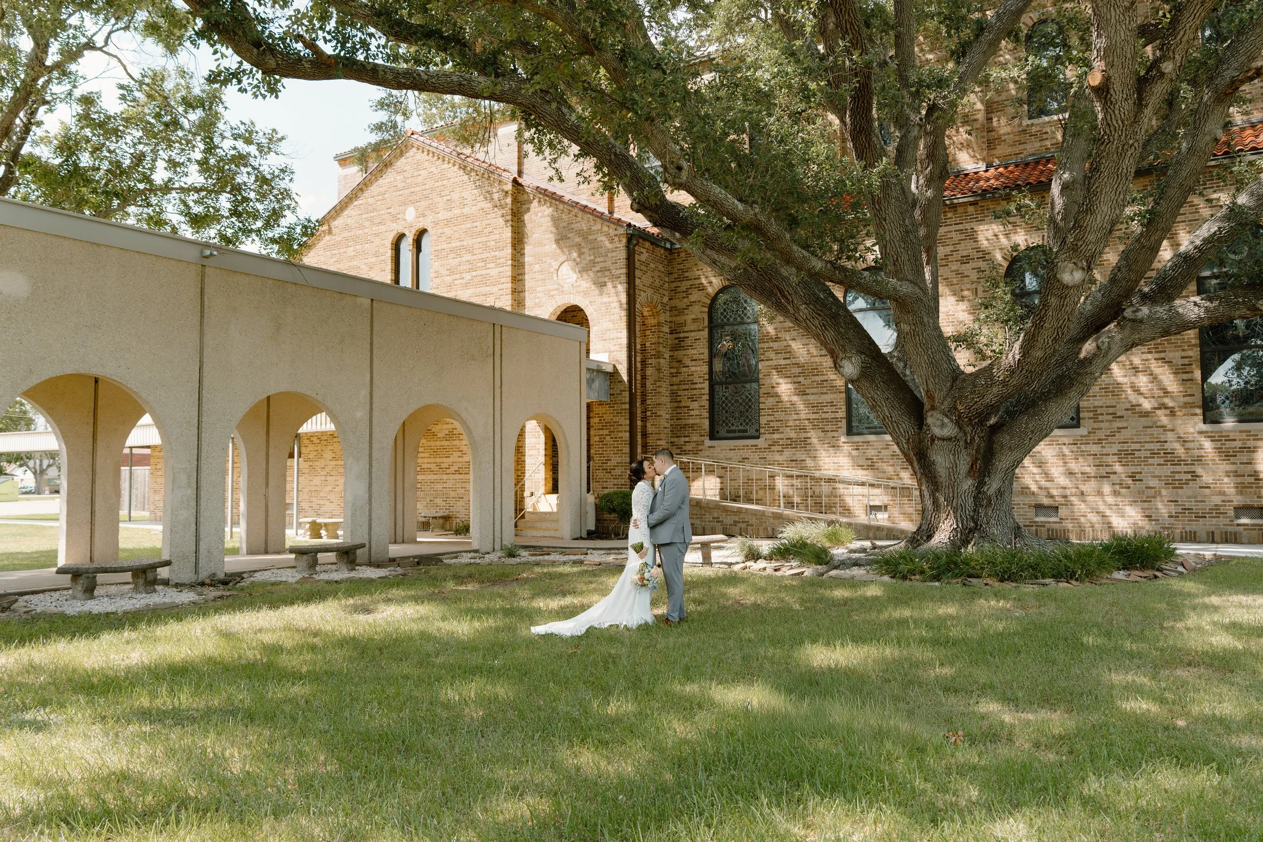 A bride and groom standing close under a large tree with a brick church in the background, during a wedding outside.