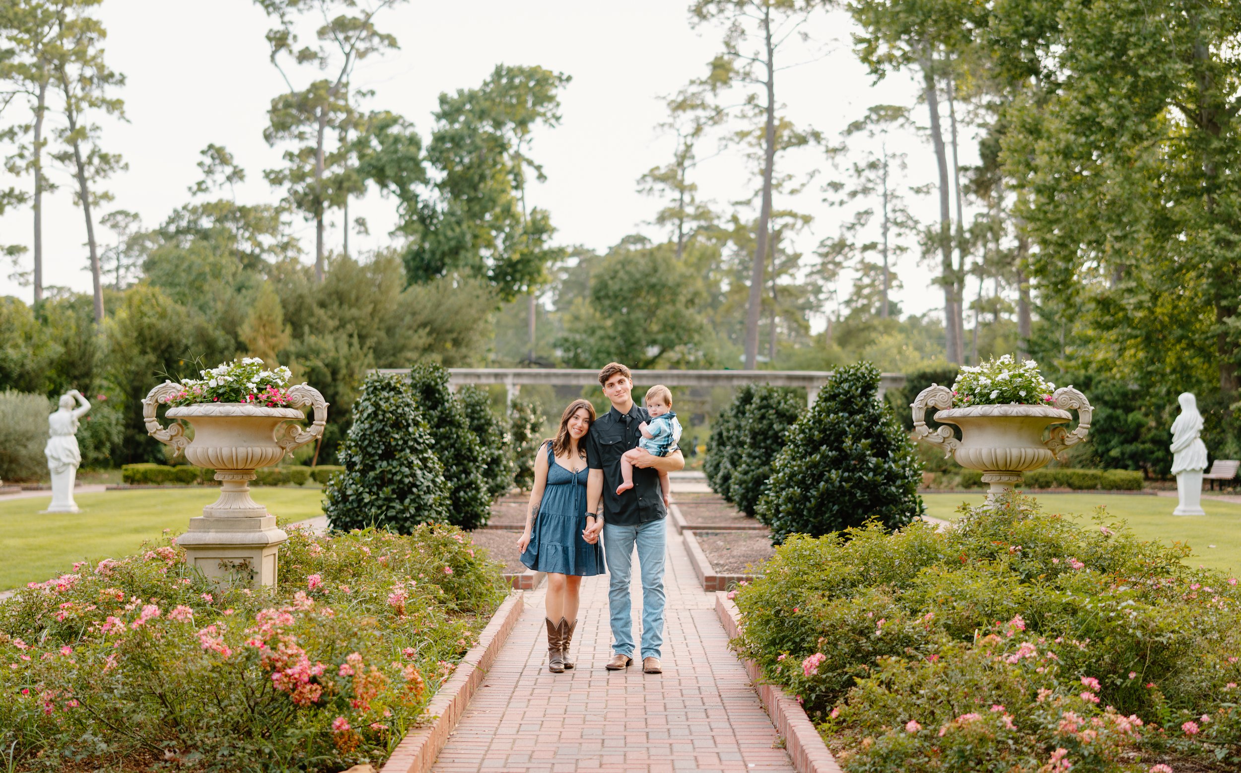 A family of three walking hand in hand through a beautifully maintained garden with large floral pots, statues, and green trees in the background.