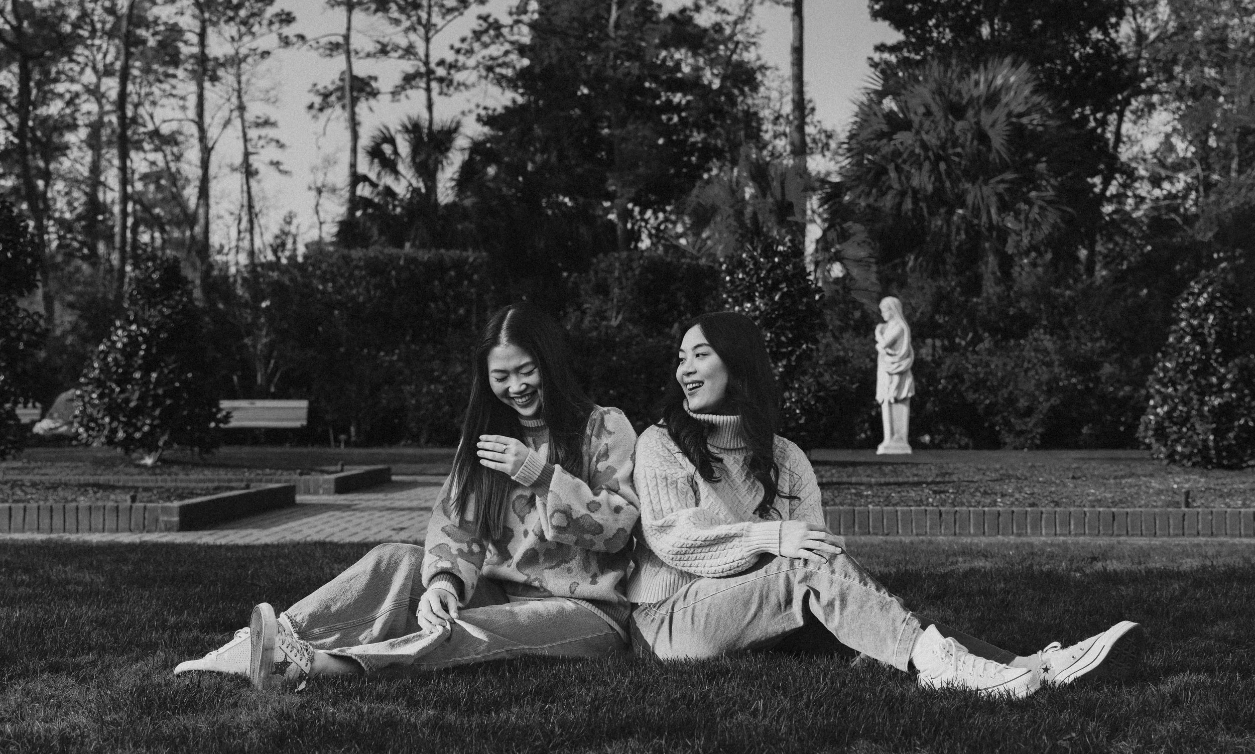 Two women sitting on grass in a park, laughing and smiling, with trees, bushes, and a statue in the background, in black and white.