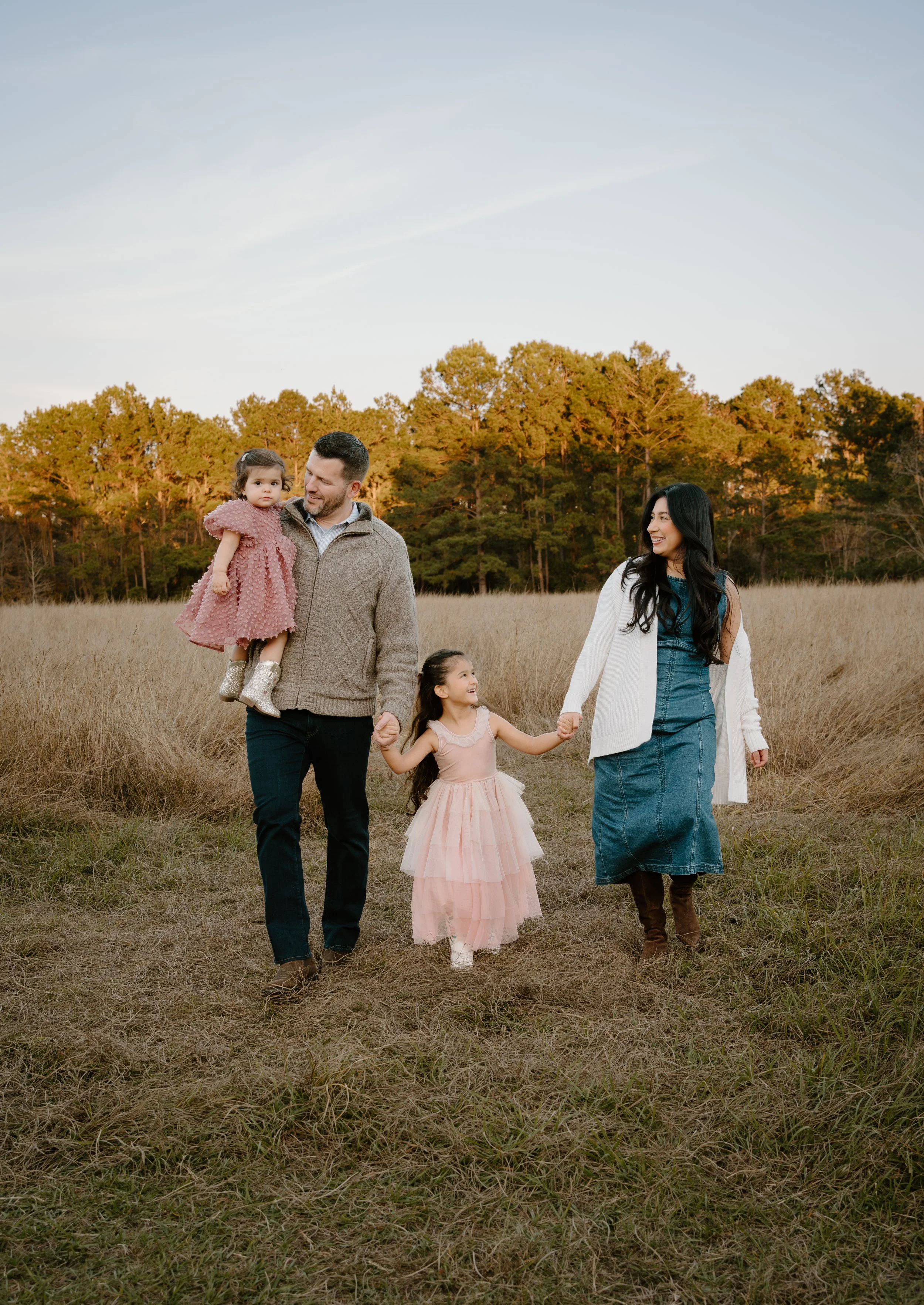 A family of four walking through a grassy field in the evening, holding hands and smiling, with trees in the background.