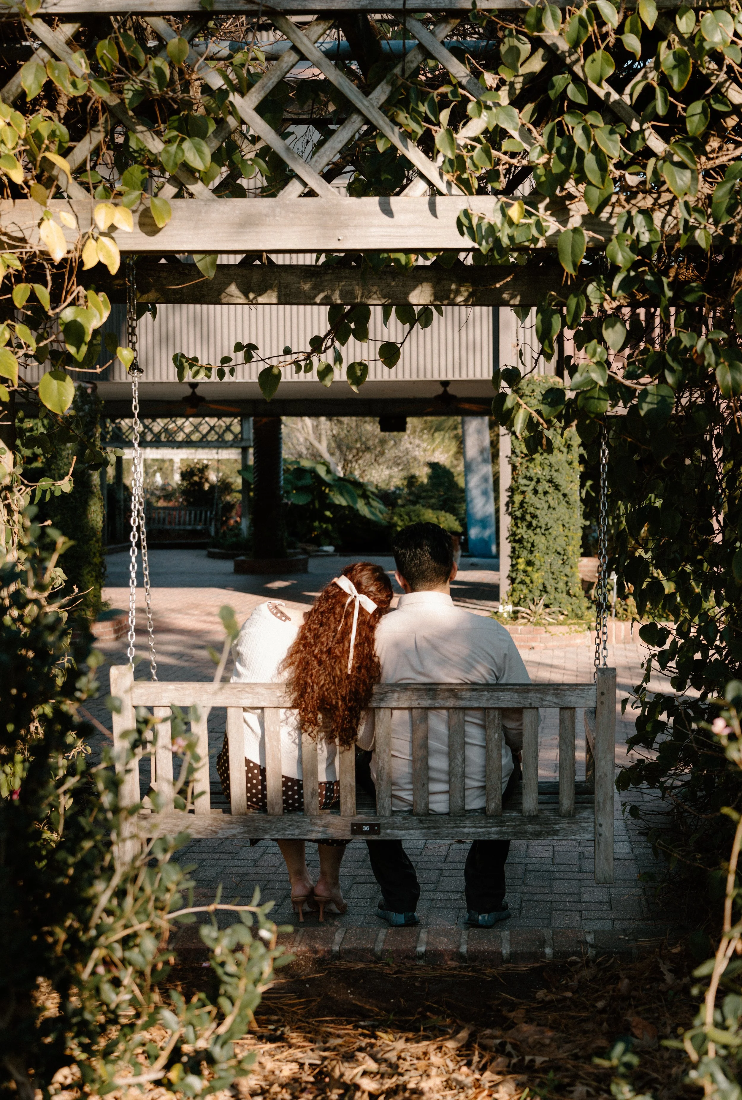 A couple sitting on a wooden swing bench under a trellis, sharing a quiet moment, surrounded by greenery.