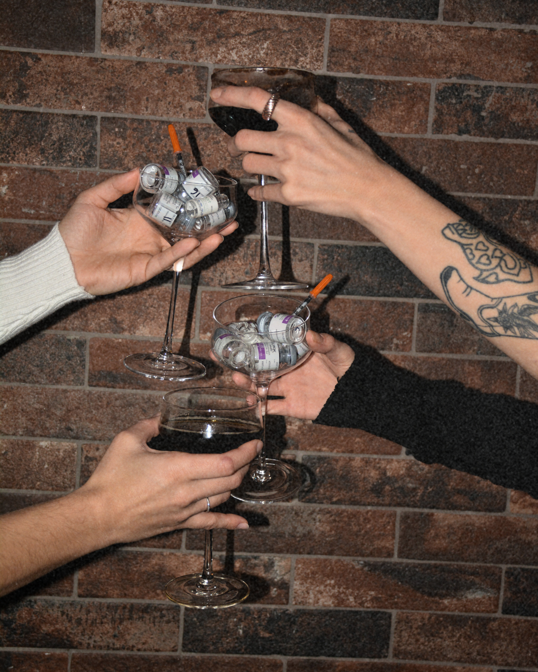 People holding wine glasses filled with syringes and vials, raising a toast against a brick wall background.