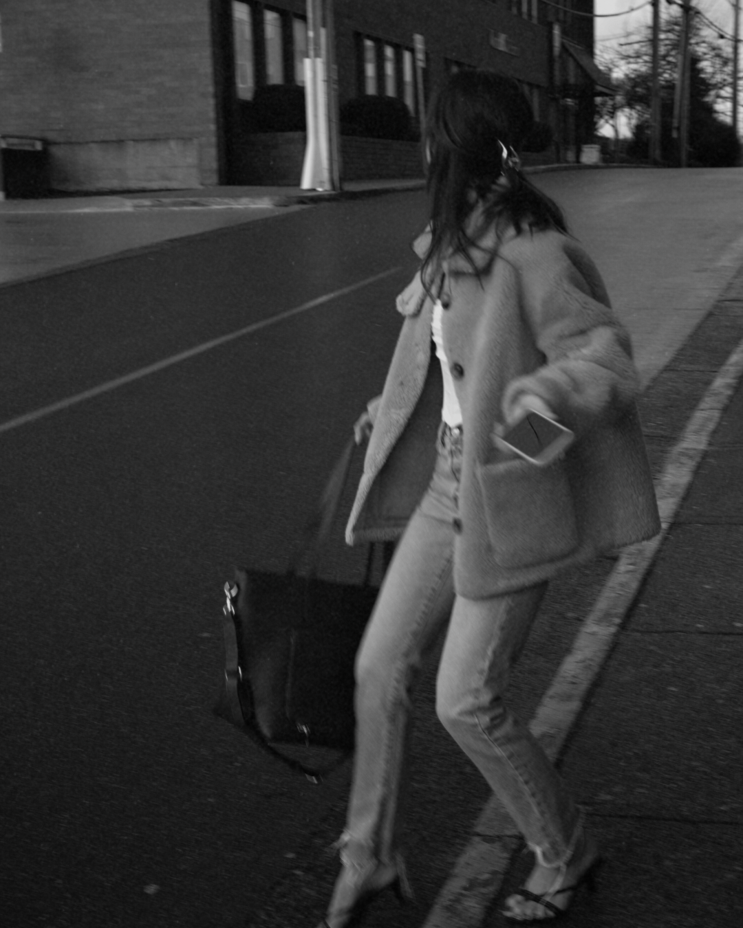A woman walking on the street at dusk, pulling a rolling suitcase, wearing a coat and high heels, holding a smartphone in her right hand.