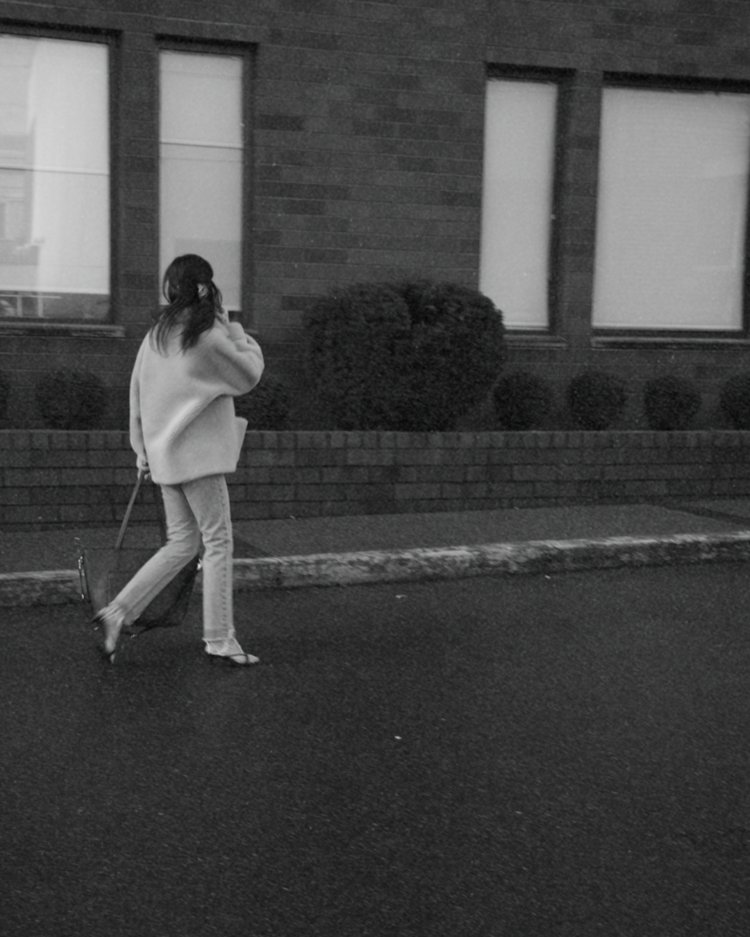 A woman wearing a coat, pants, and sandals walking on the sidewalk while holding a bag and phone, in front of a building with three vertically aligned windows and shrubbery.