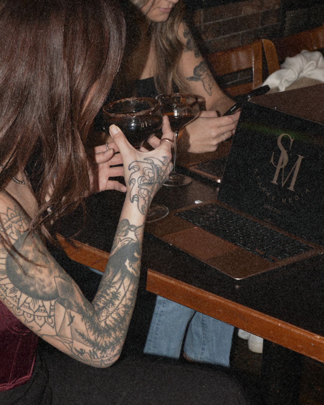 Two women with tattoos sitting at a bar, holding glasses of dark beverage, with a laptop open in front of them.