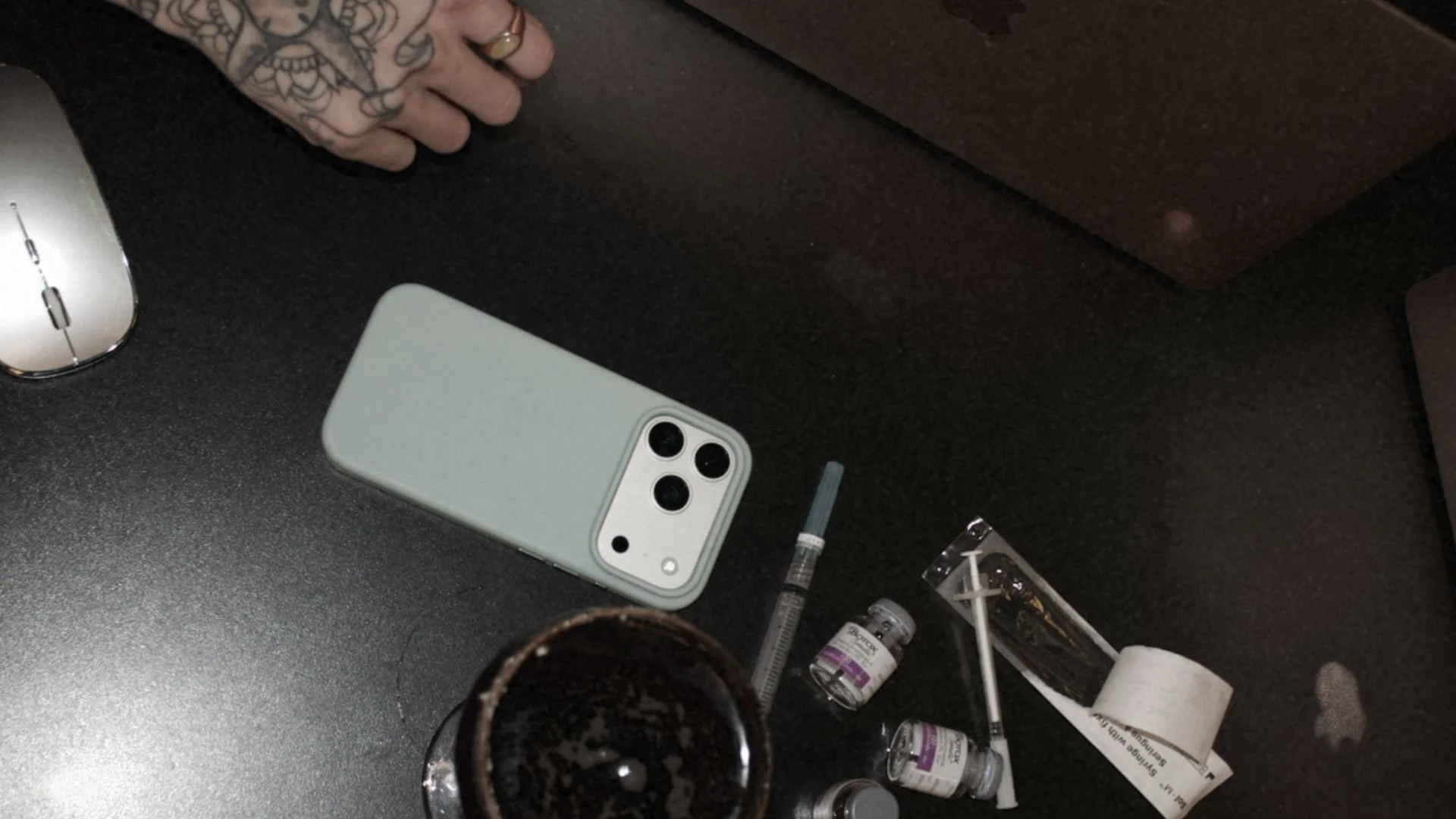 Top view of a black desk with a white smartphone, a white computer mouse, a glass of soda, syringes, small vials, and a small packet.