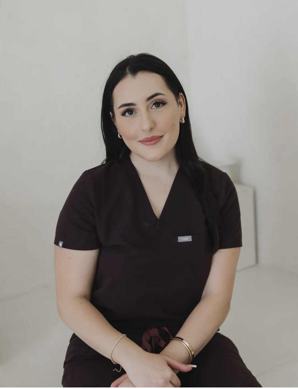 A young woman with black hair, wearing a brown uniform with a name tag labeled 'FIGS', sitting against a plain white wall, smiling gently at the camera.