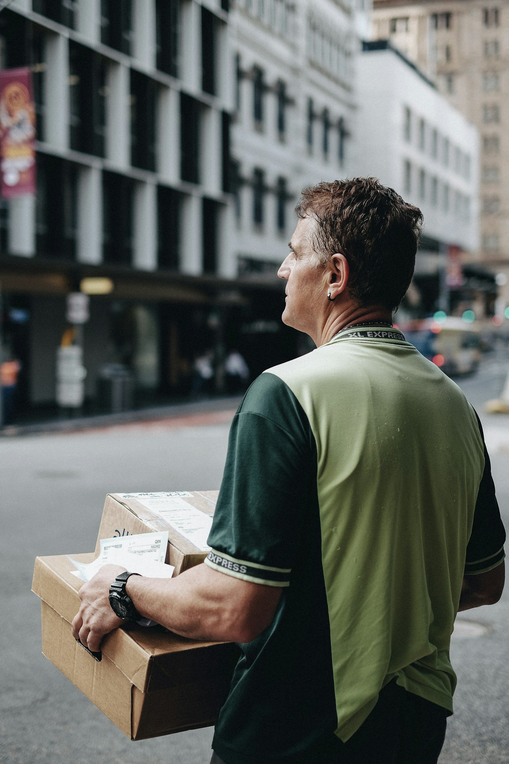 A delivery person holding a cardboard box and some papers on a city sidewalk, wearing a green and black uniform.