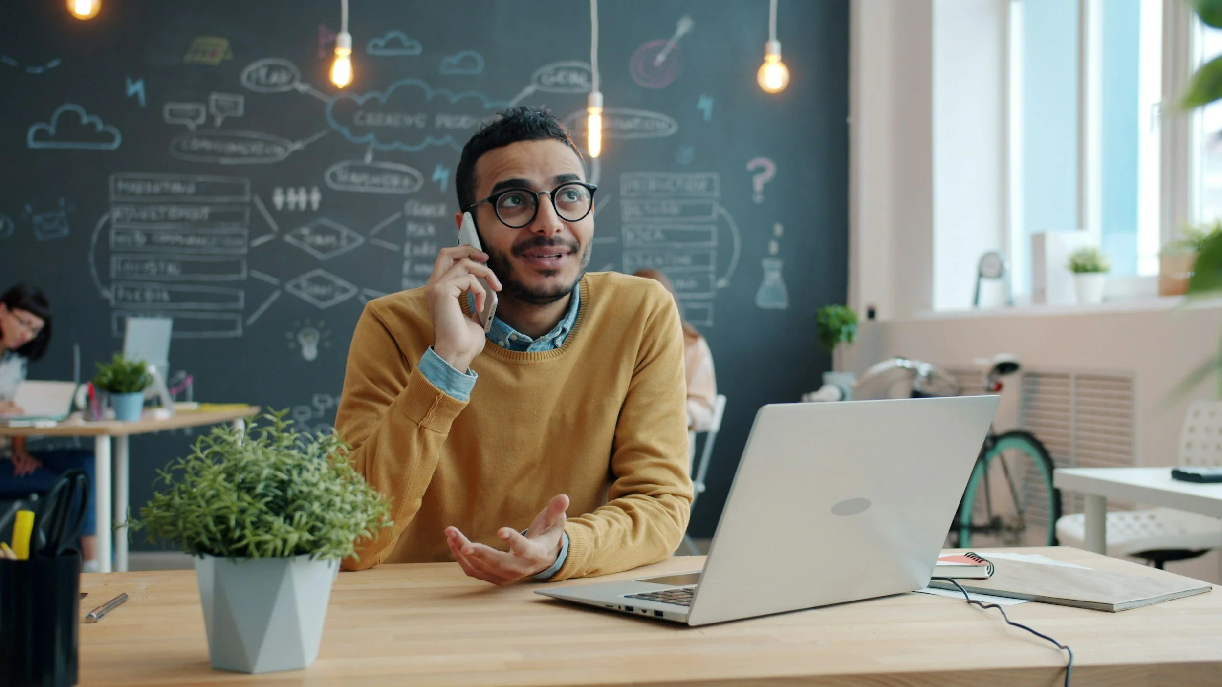 A man in glasses and a mustard sweater talking on his phone at his desk, with a laptop, notebooks, and a potted plant in front of a blackboard with drawings and notes.