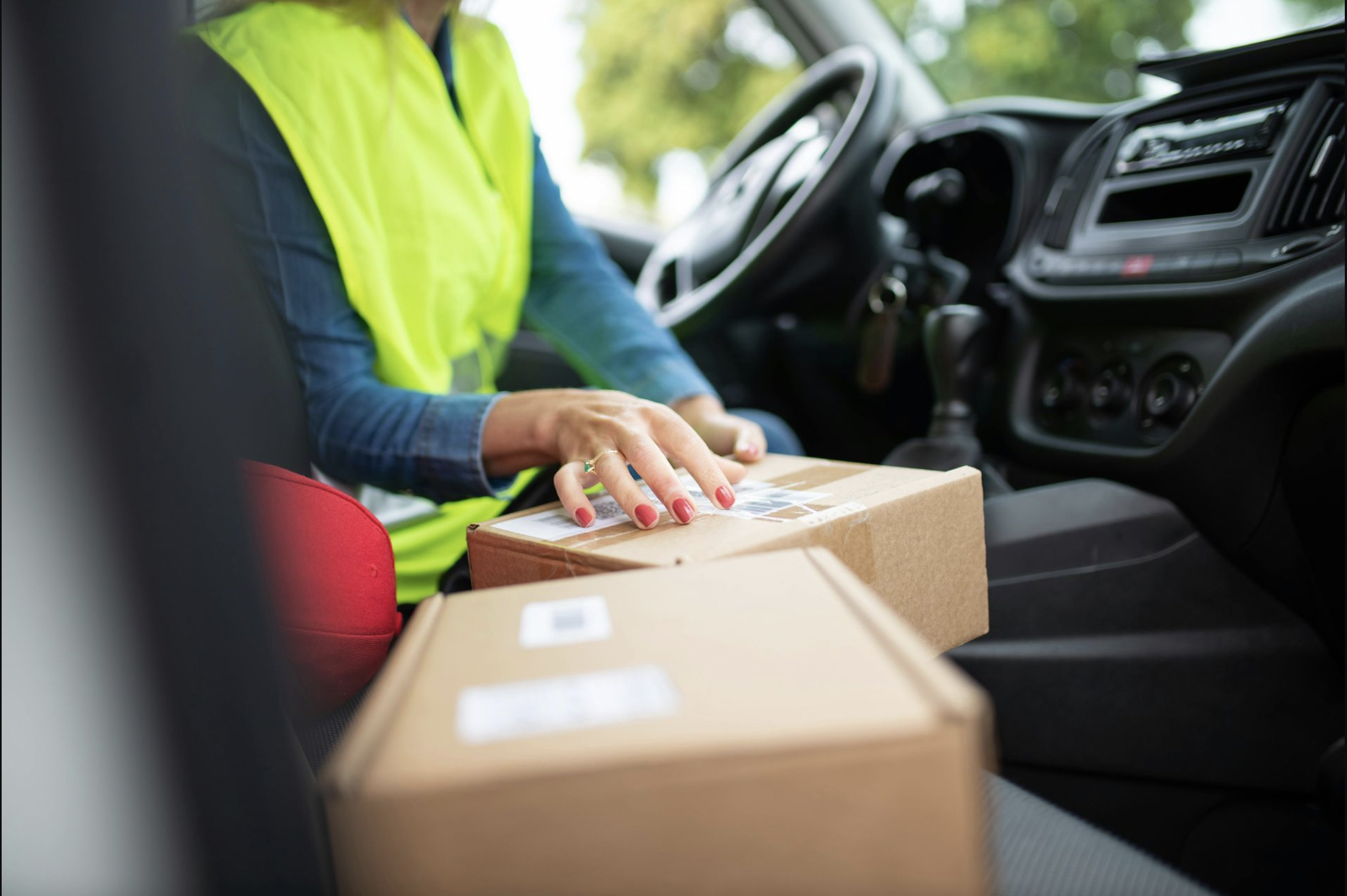 A person in a yellow safety vest sitting in the driver's seat of a vehicle, handling a cardboard box, with a second box in the foreground.