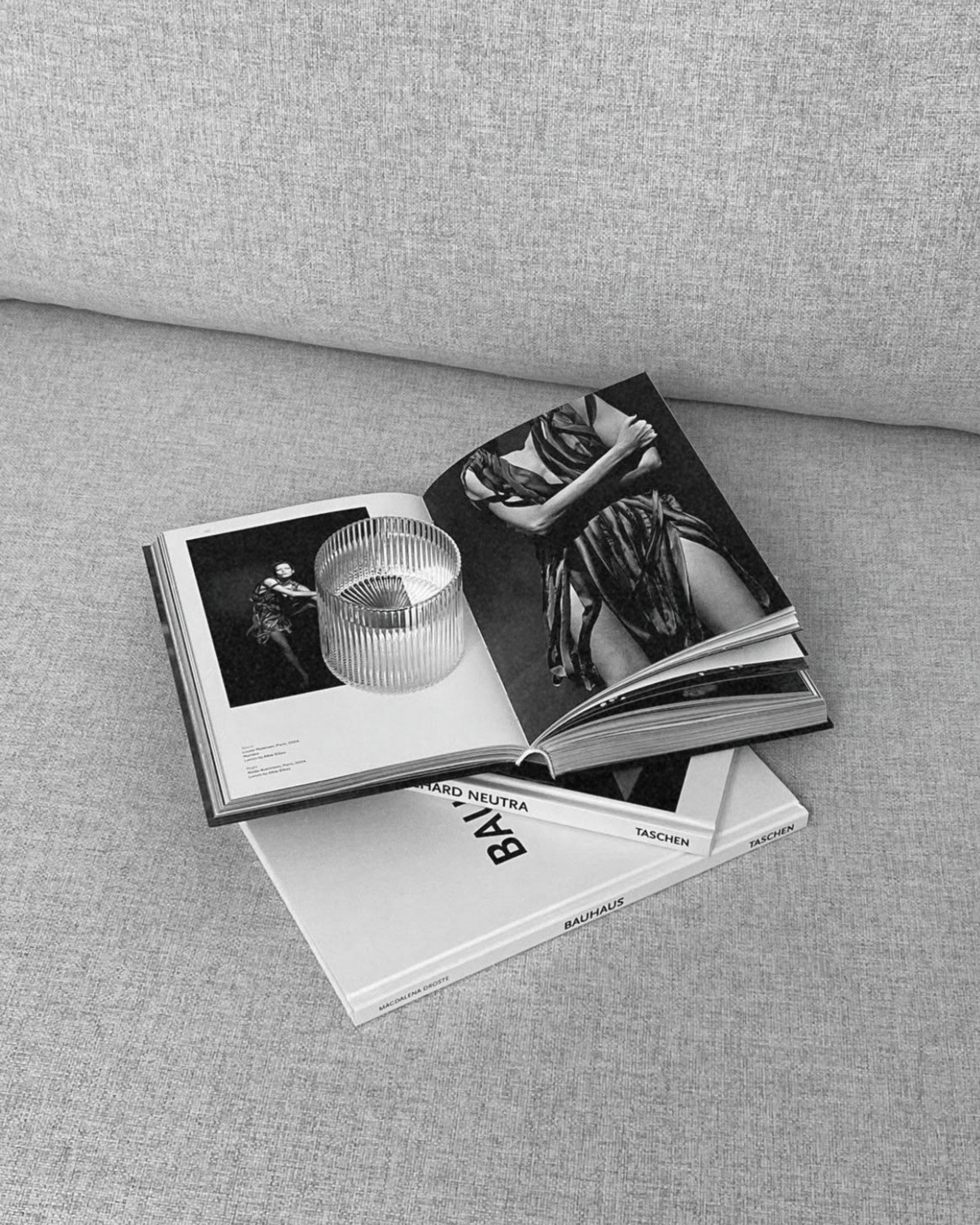 An open magazine featuring a black-and-white fashion photo with a transparent, ribbed glass object on top, resting on top of two other closed books on a light-colored fabric surface.