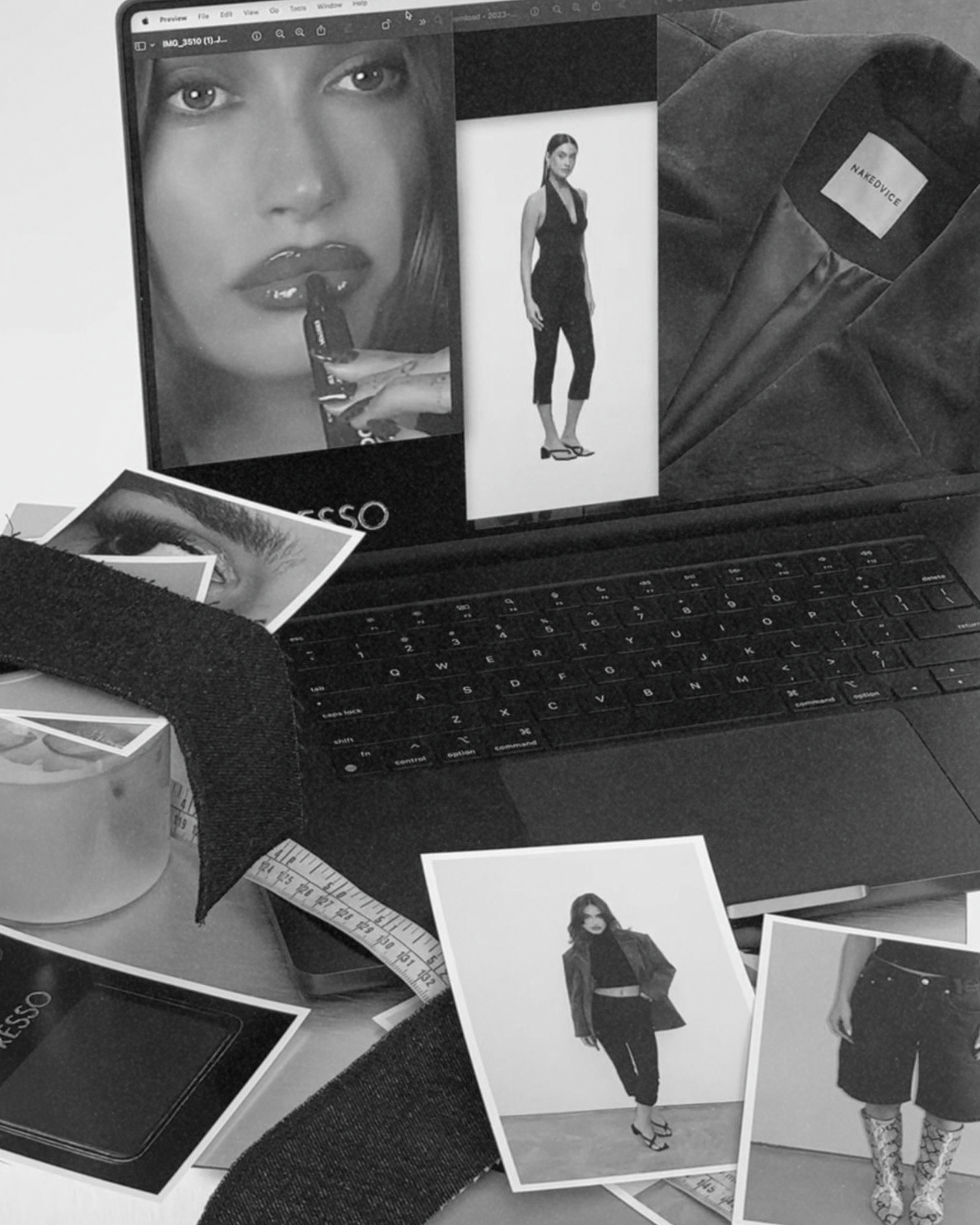 A workspace with a laptop displaying fashion photos, surrounded by printed fashion photographs, a measuring tape, and a patch labeled 'NAKEDVICE'. The photos include close-ups of women's faces, full-body images of women in styles like jumpsuits, blouses, and skirts, with some models wearing high heels and boots.