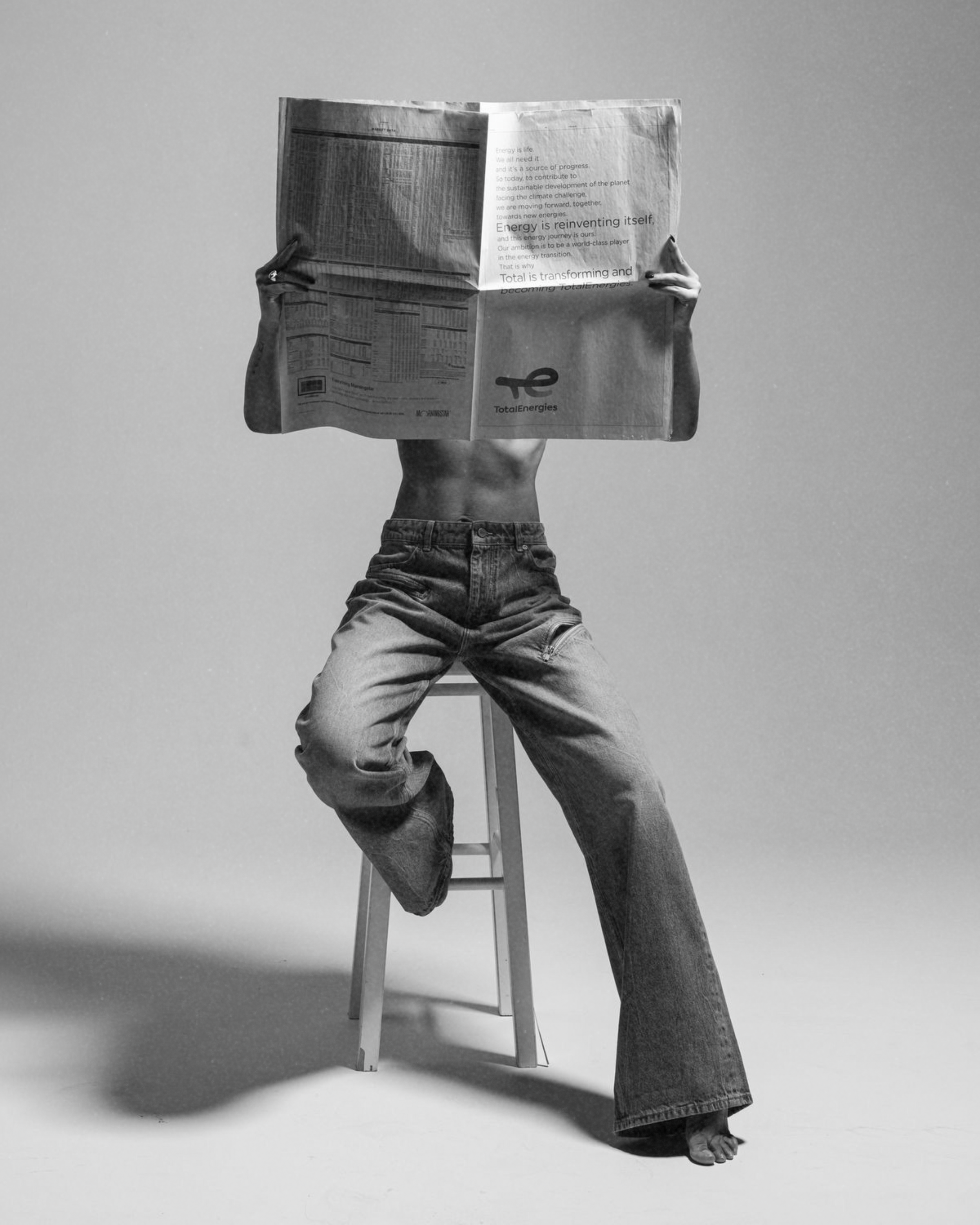 Person sitting on a stool, holding a large newspaper in front of their face, with only their torso and legs visible.