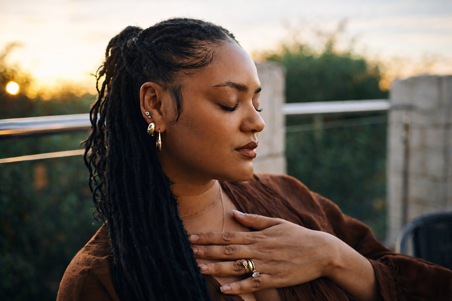 A woman with dreadlocks and gold jewelry sits outdoors with her eyes closed and hand on her chest at sunset.