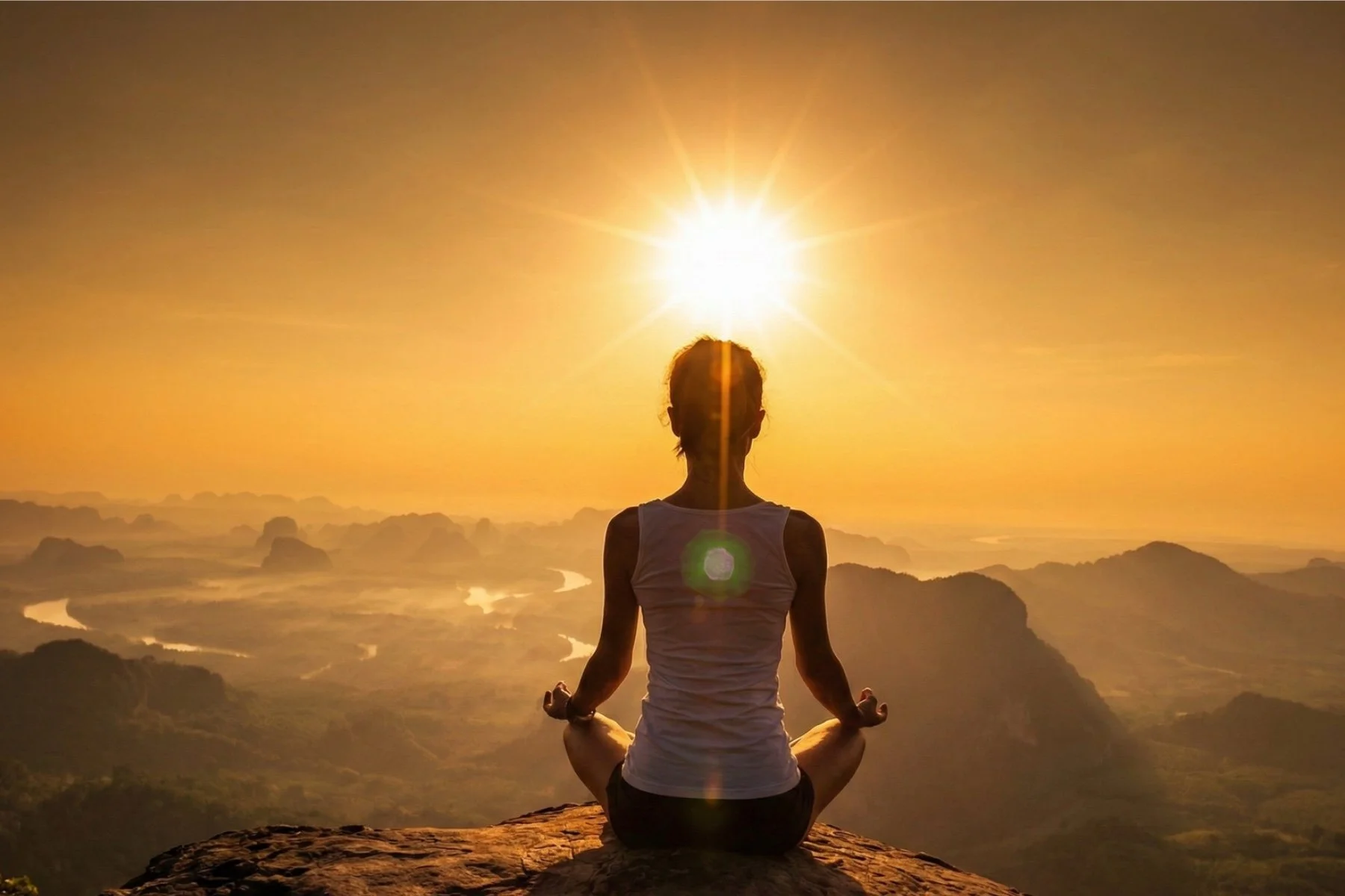 A person practicing yoga in a seated meditation pose on a rocky ledge at sunset with mountains and winding rivers in the background, with the sun directly behind them.